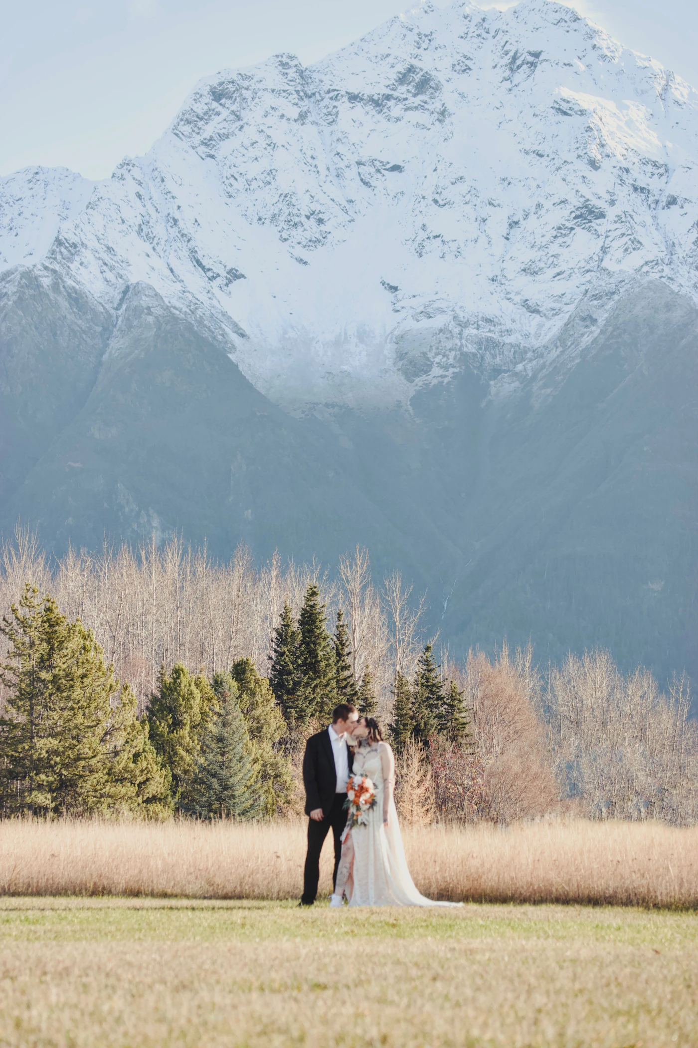A bride and groom sharing a romantic first kiss in a flower-filled alpine meadow with snow-capped mountain peaks visible in the background at their mountain wilderness wedding
