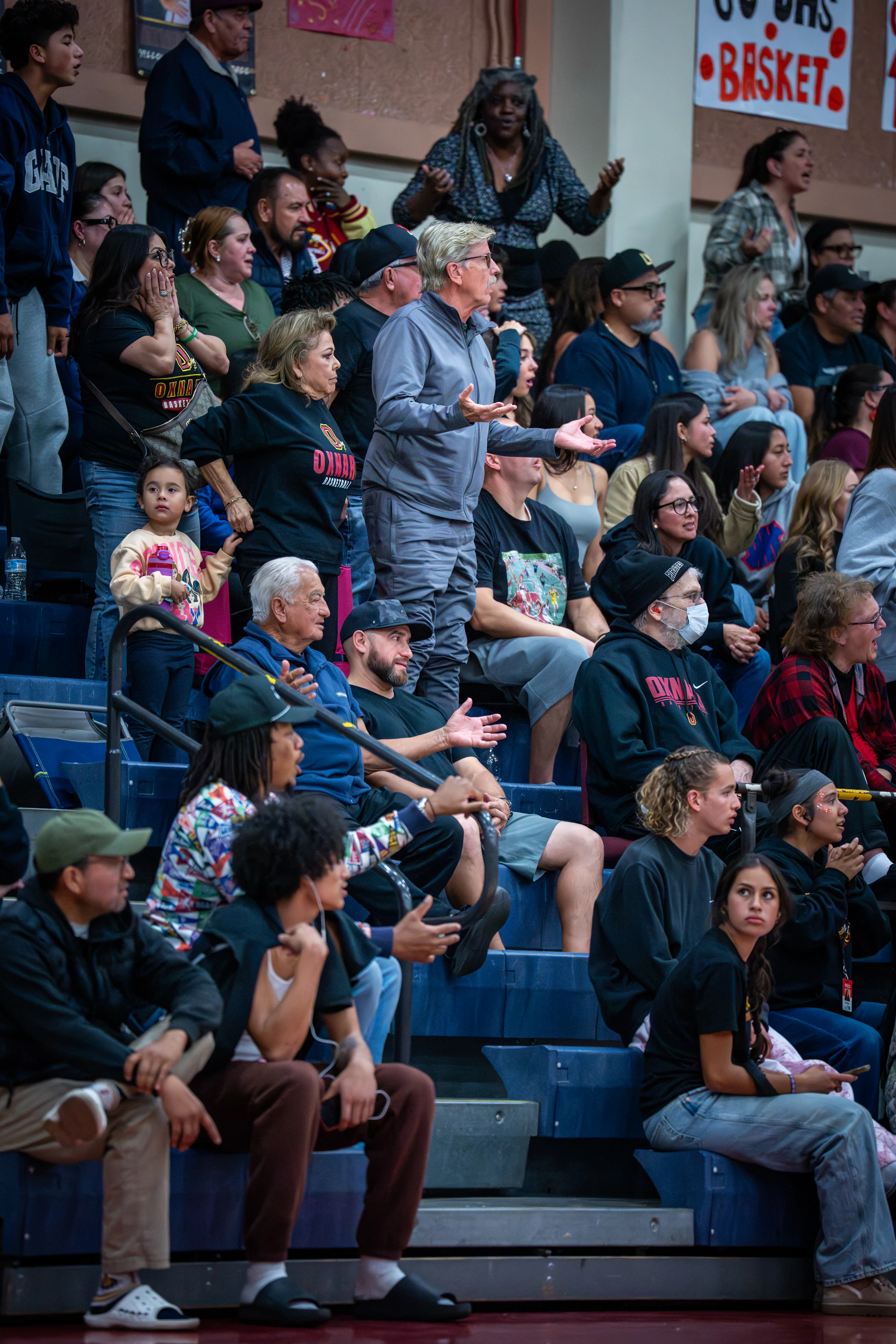 A packed set of gym bleachers at a school game, with one fan on his feet holding his arms out at a call
