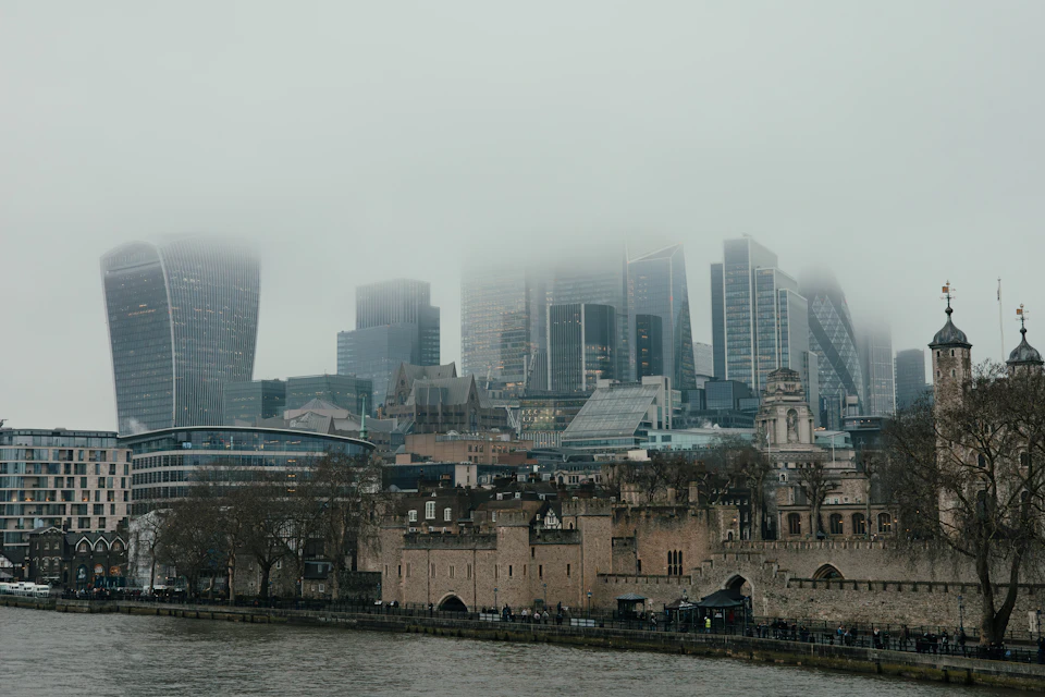 The Tower of London on the Thames under low grey cloud, with the City's skyscrapers, including the Walkie-Talkie, fading into the mist behind bare winter trees