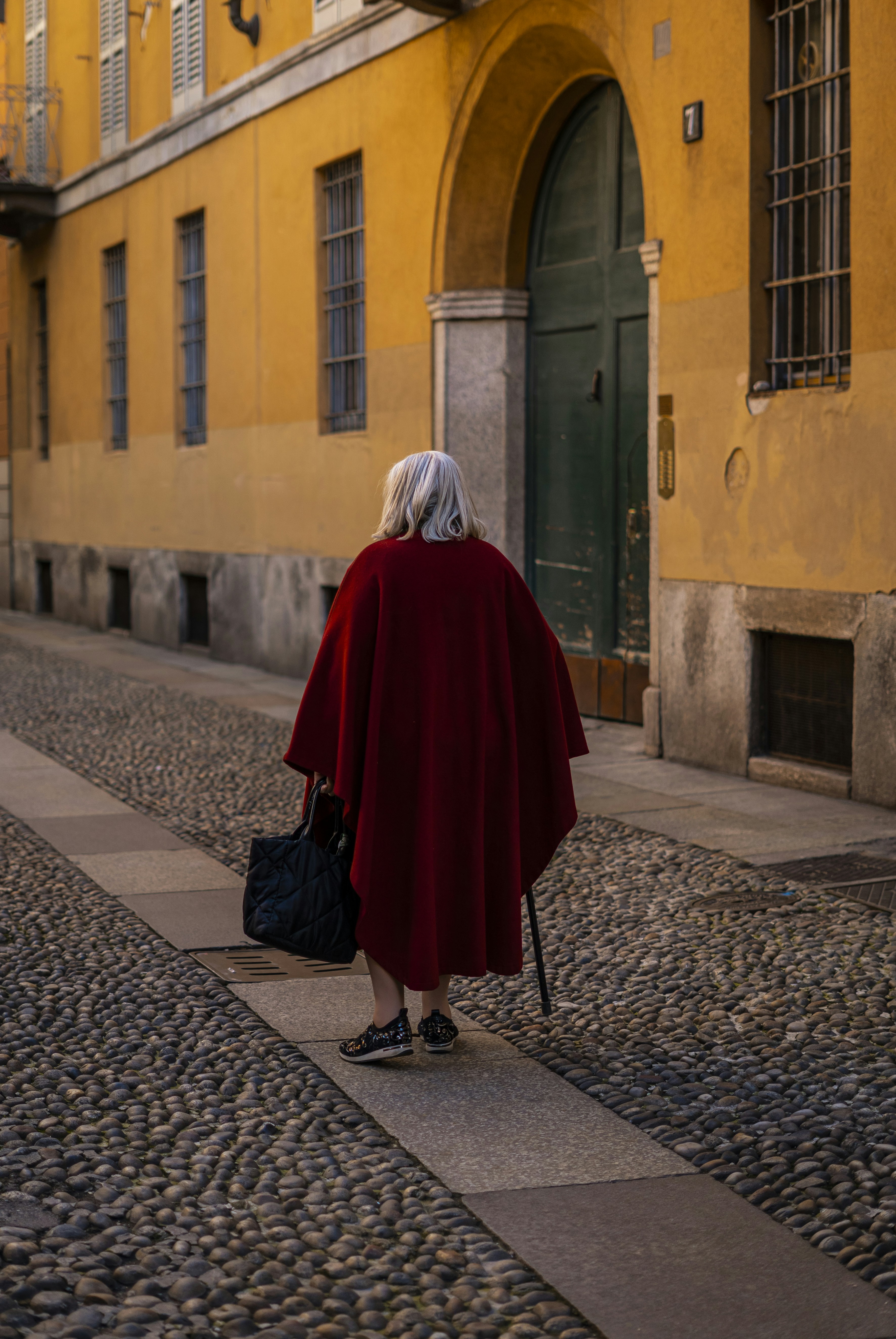 Elderly person in red cape walks down cobblestone street.