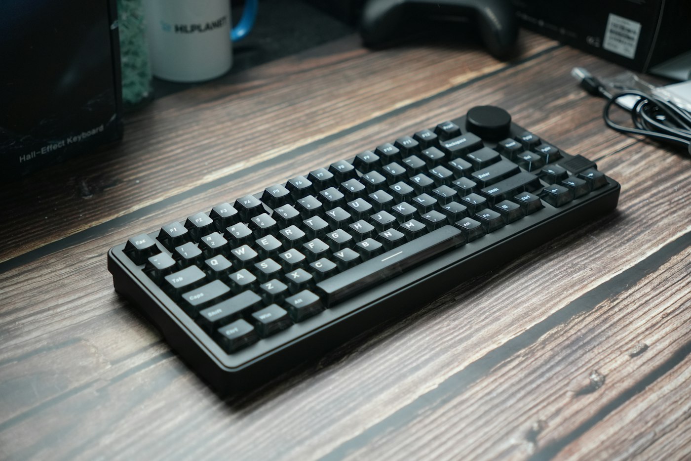 A black mechanical keyboard with a volume knob on a wooden desk beside a mug and cables