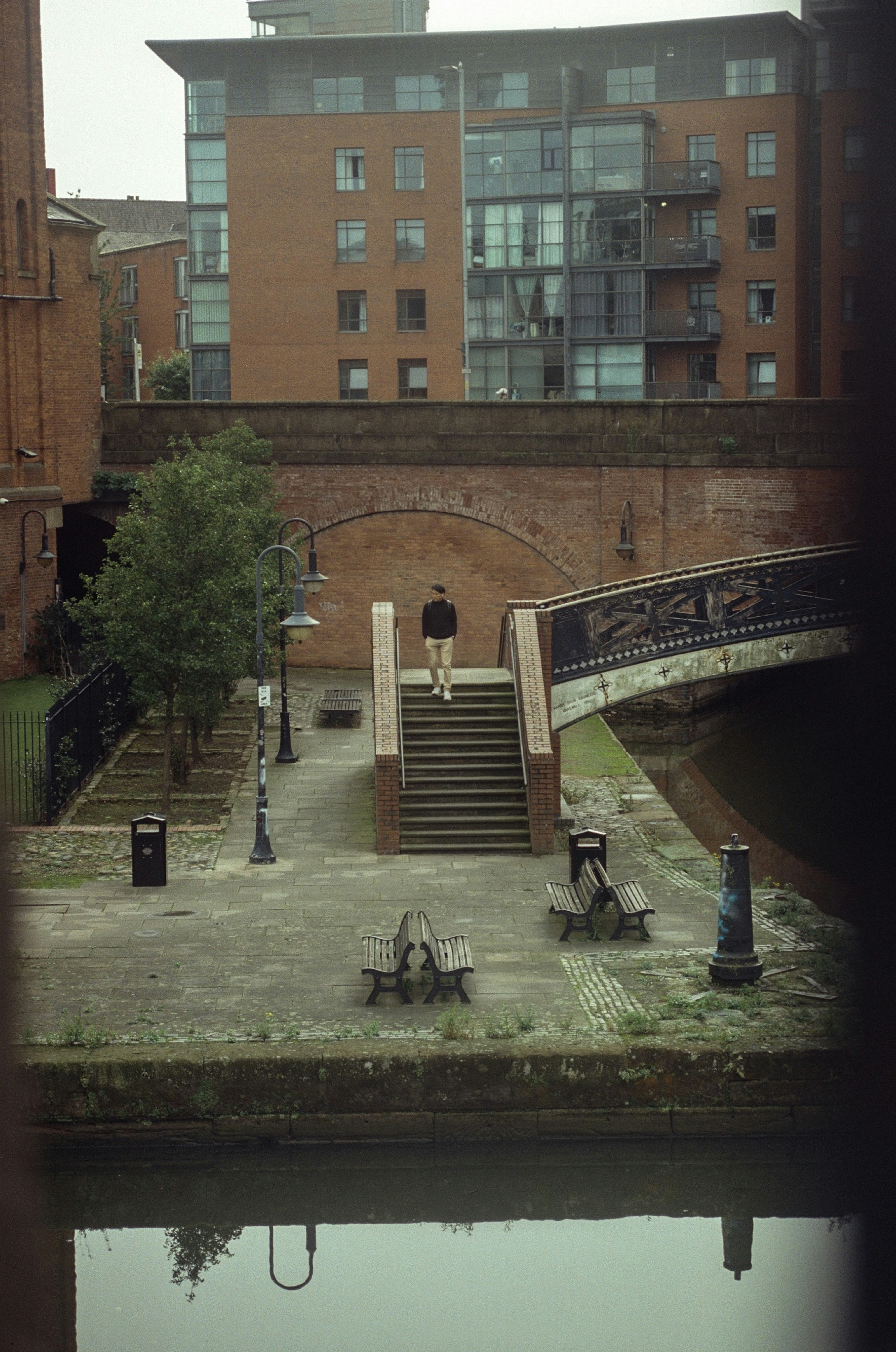 Man standing on stairs by canal with canal and bridge.