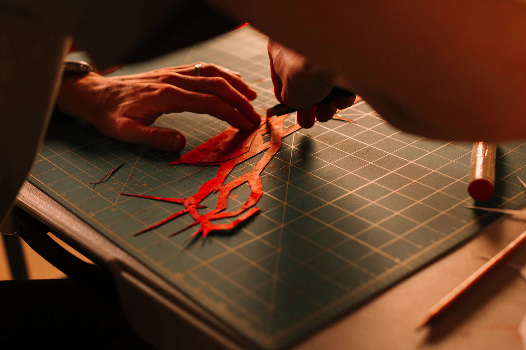 Two hands cutting an intricate red paper shape with a knife on a gridded cutting mat