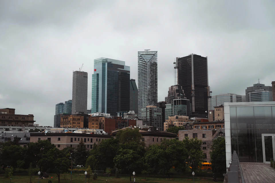 The image depicts a cityscape with tall buildings, including skyscrapers and shorter structures, lining both sides of the frame. The tallest building on the left side is partially obscured by clouds, while the tallest building on the right side stands out against the sky. In the foreground, there are trees providing a touch of nature amidst the urban landscape. A few cars can be seen parked along the street, and a bus is visible in the distance.