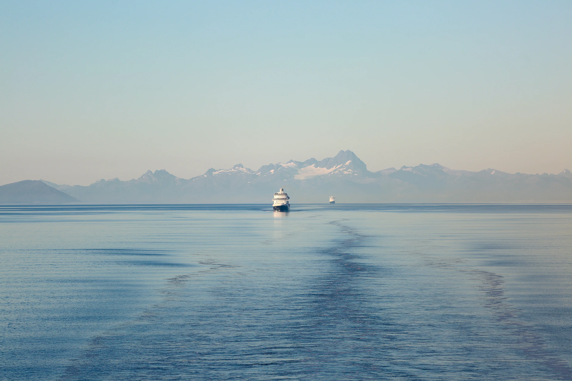 Two ships sail on a calm ocean towards mountains.