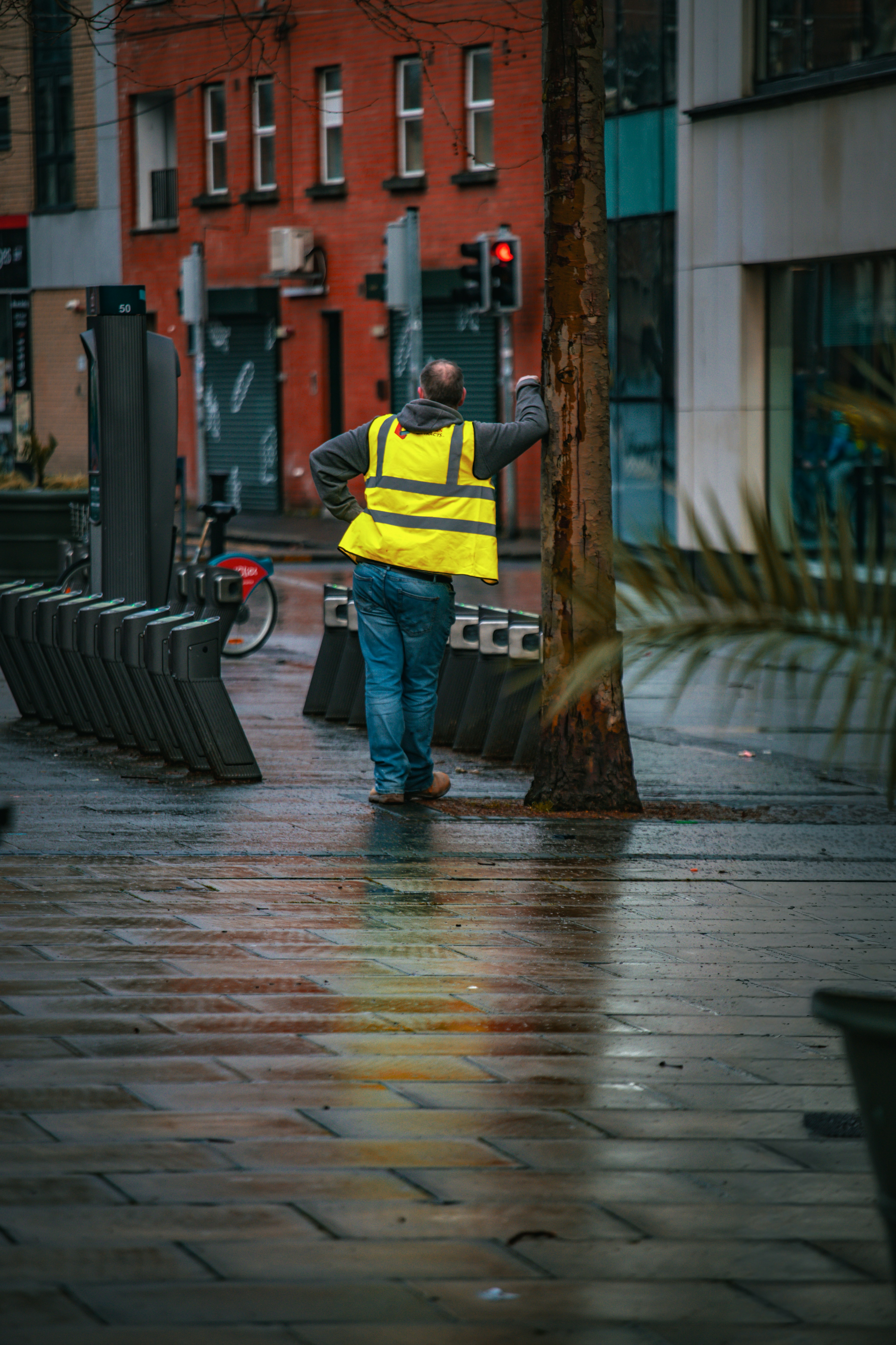 Man in yellow vest leaning against tree on wet street