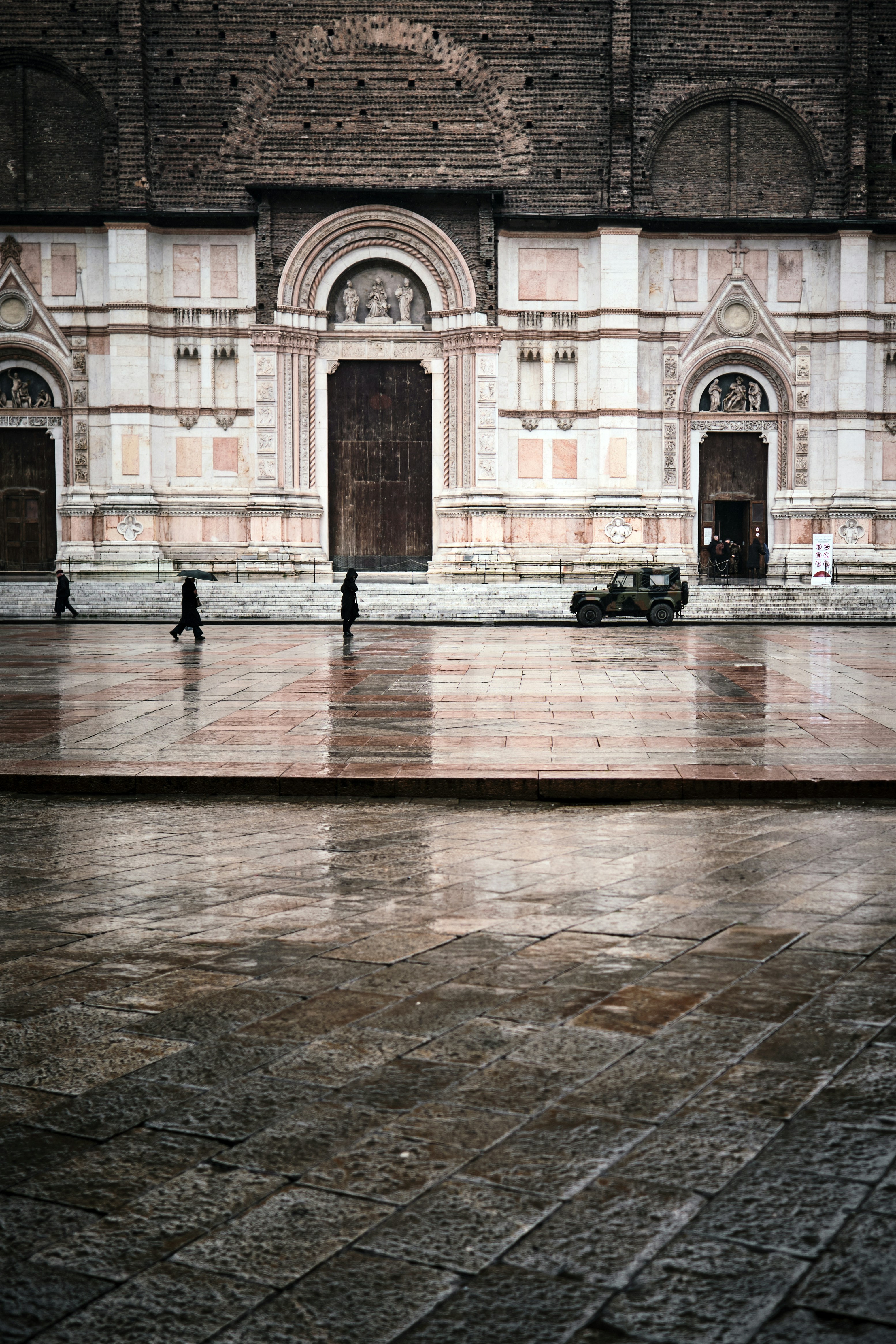 People walk across wet cobblestone plaza in front of building.