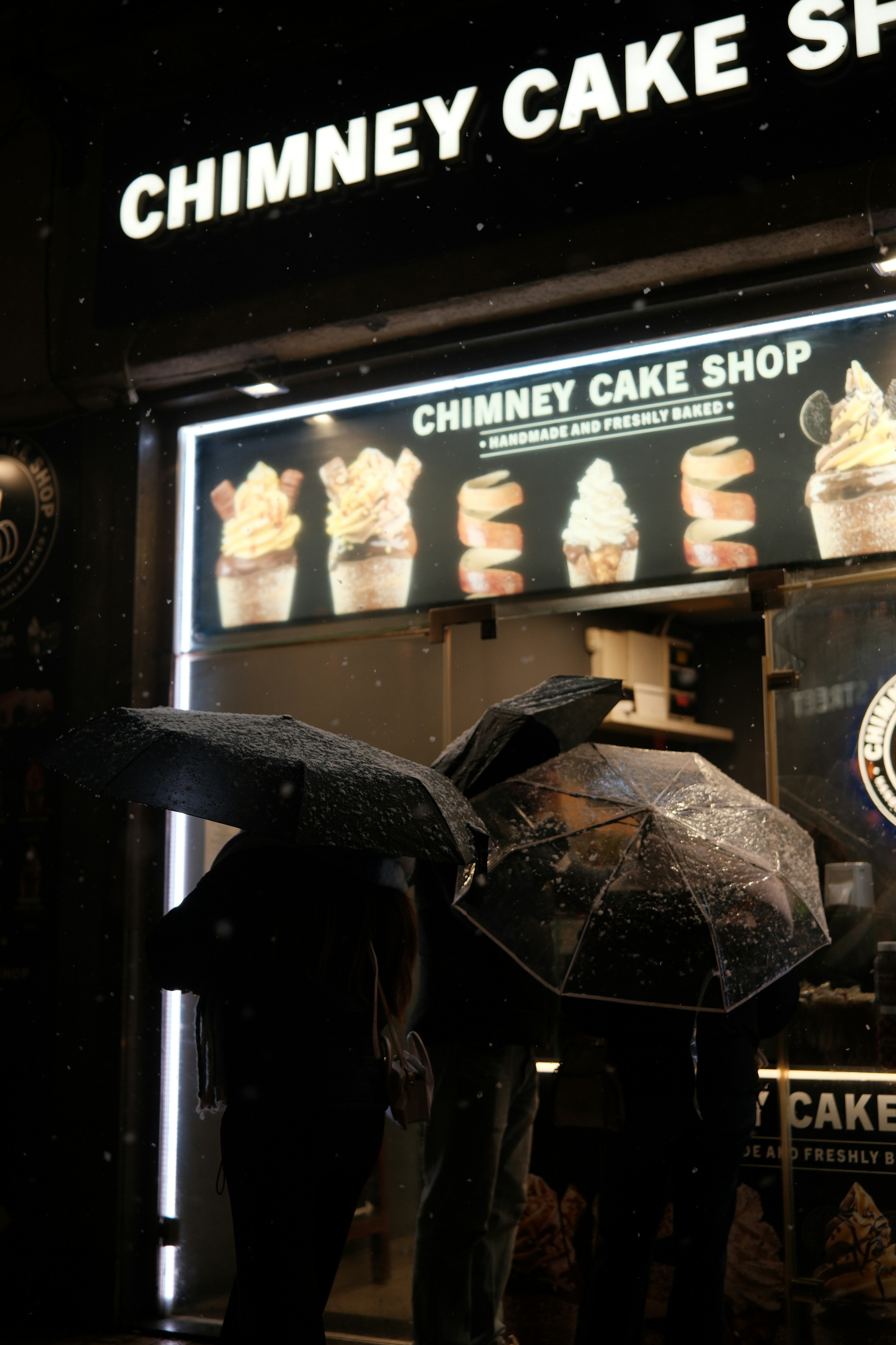 Gente con paraguas fuera de una pastelería de chimenea por la noche.