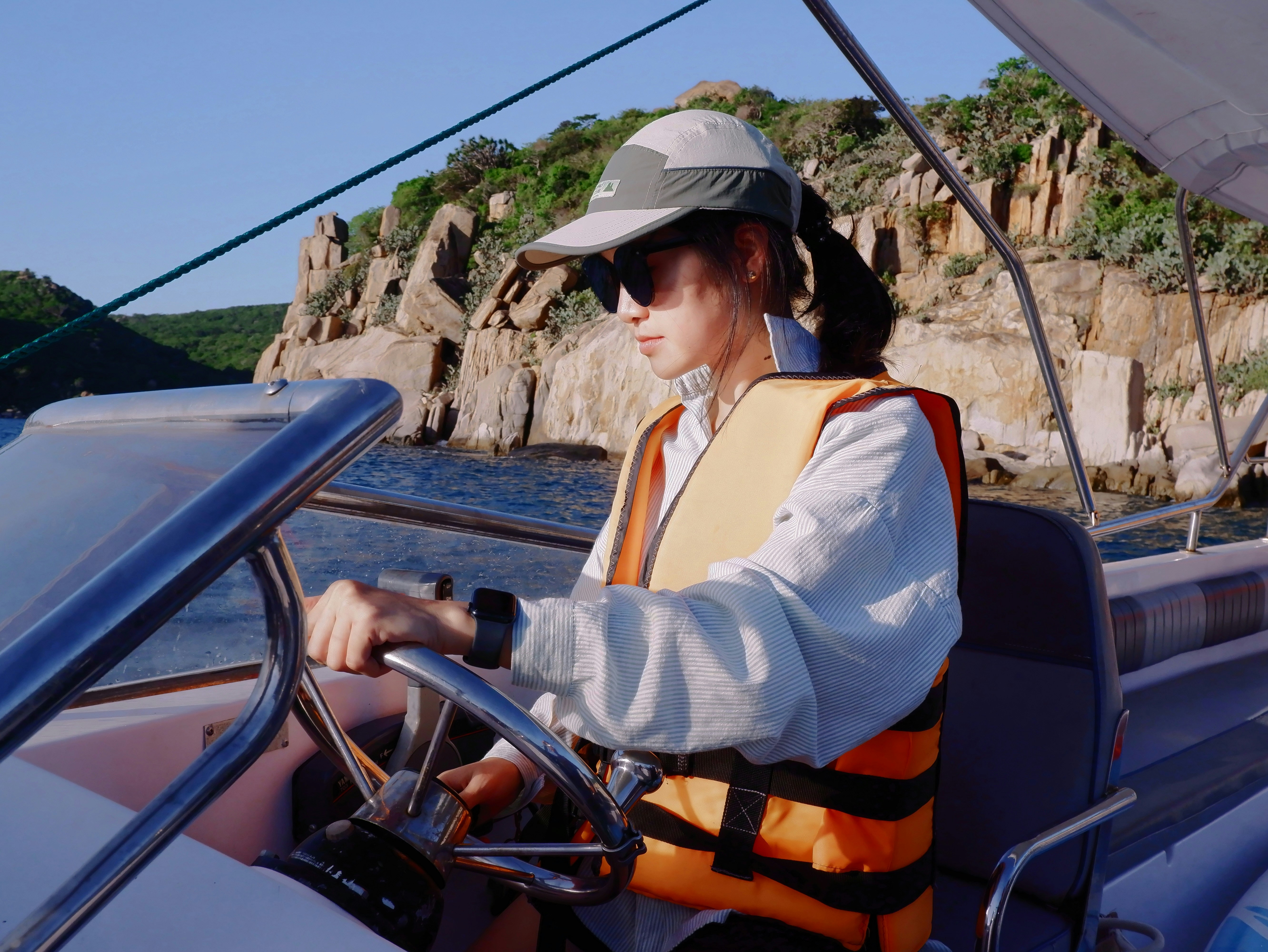 Woman in an orange life jacket and cap steering an open boat along a rocky shore, one hand on the wheel