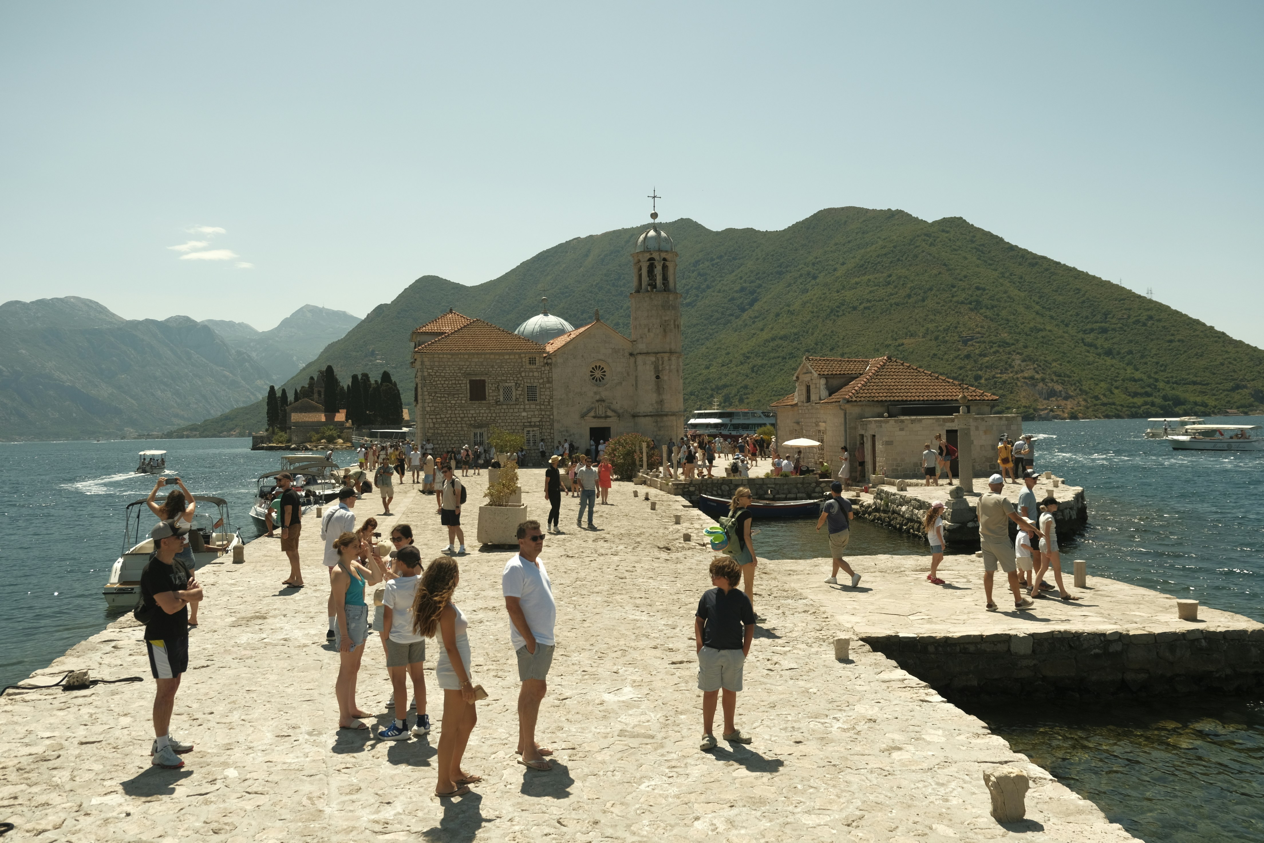 Crowds on the stone pier at Our Lady of the Rocks, the domed island church off Perast on the Bay of Kotor, the green mountains beyond — the focus of the Fašinada and Boka Night traditions
