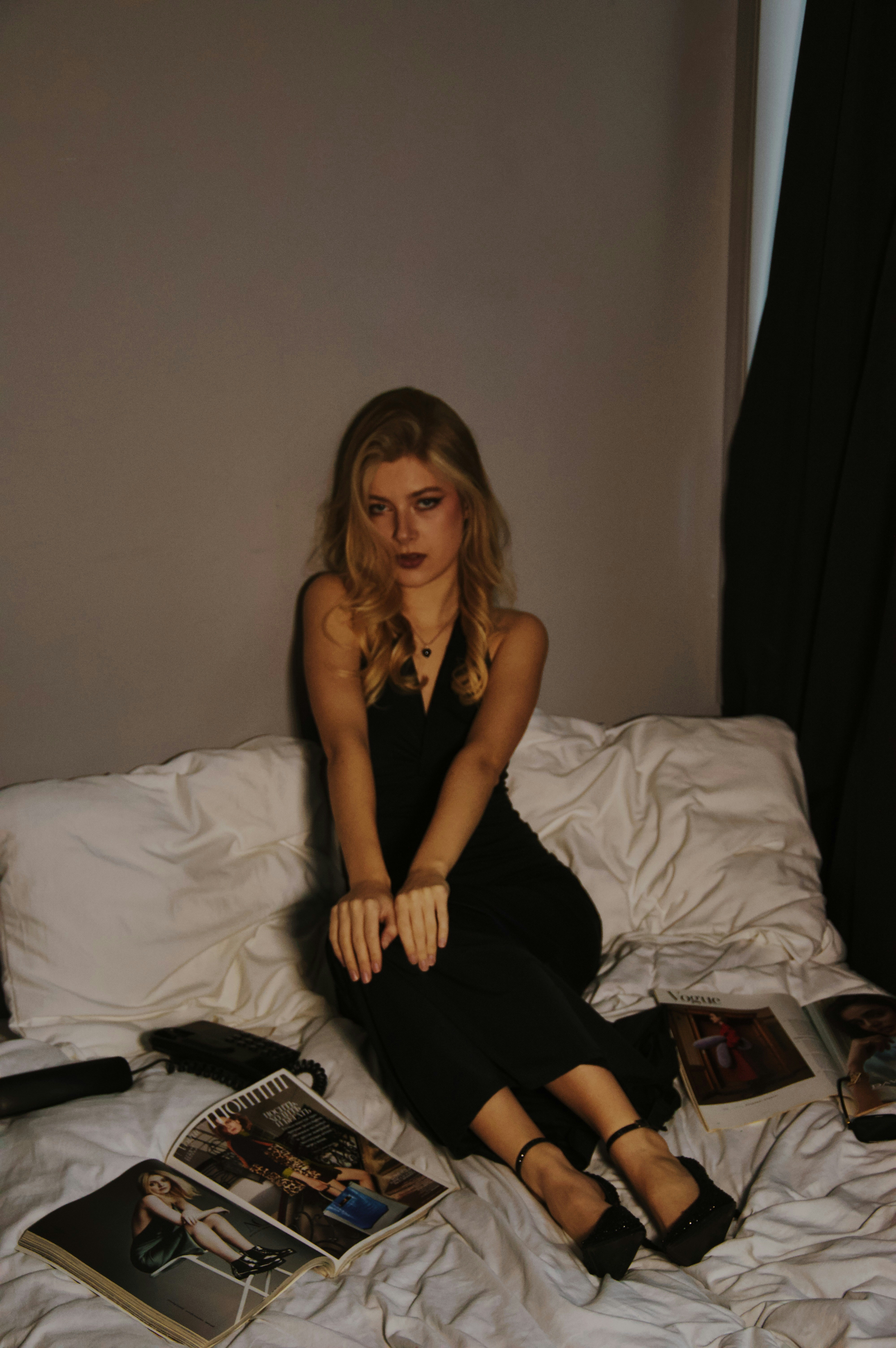 Woman in black dress sitting on a bed with magazines.
