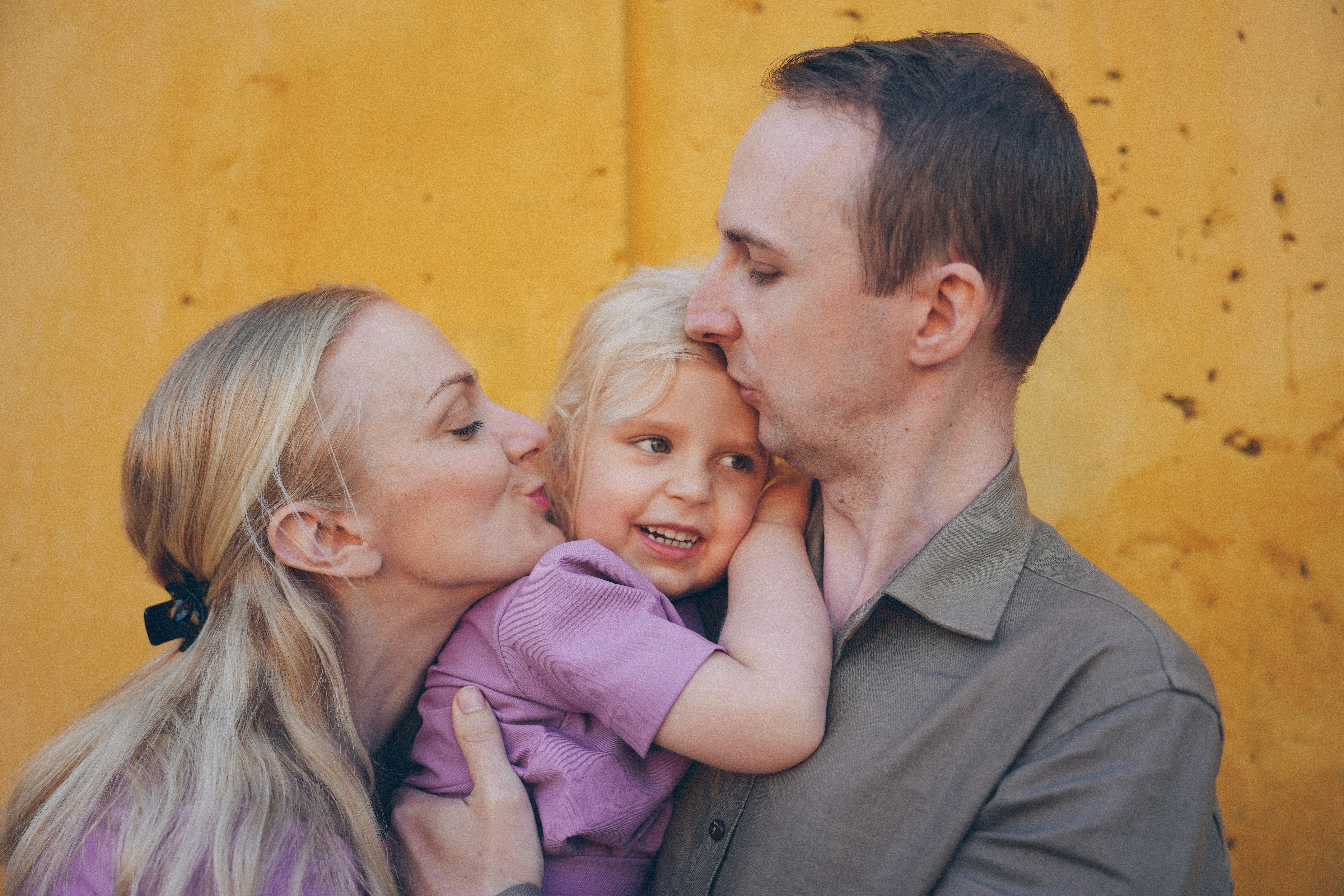 Parents kissing their daughter on a yellow wall.