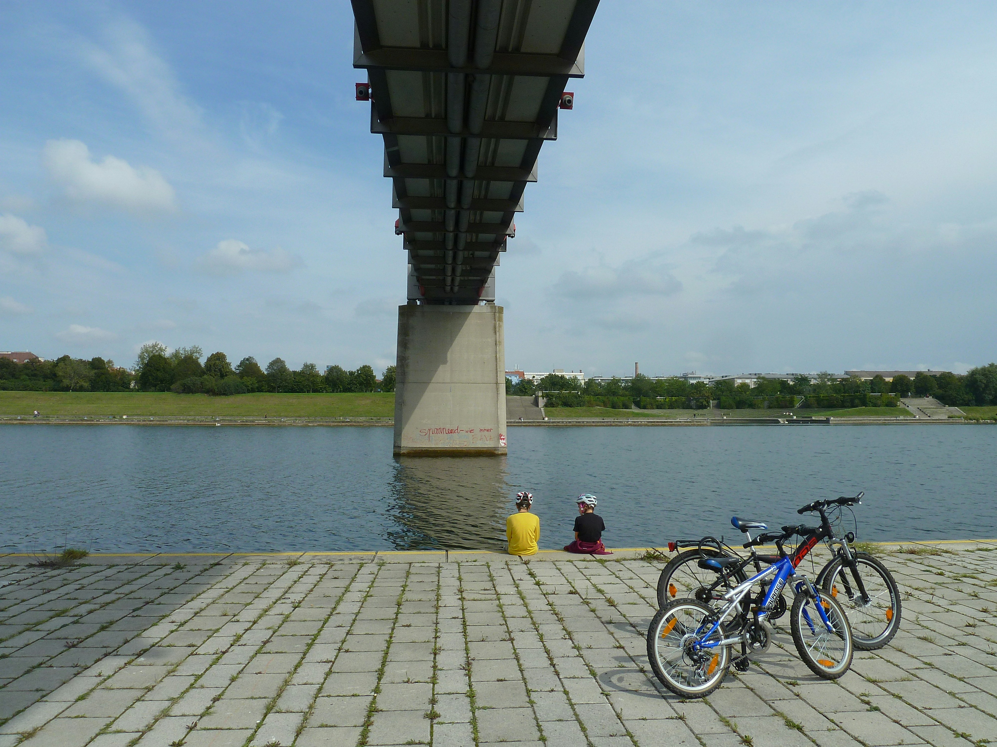 Two bicycles parked by a river under a bridge