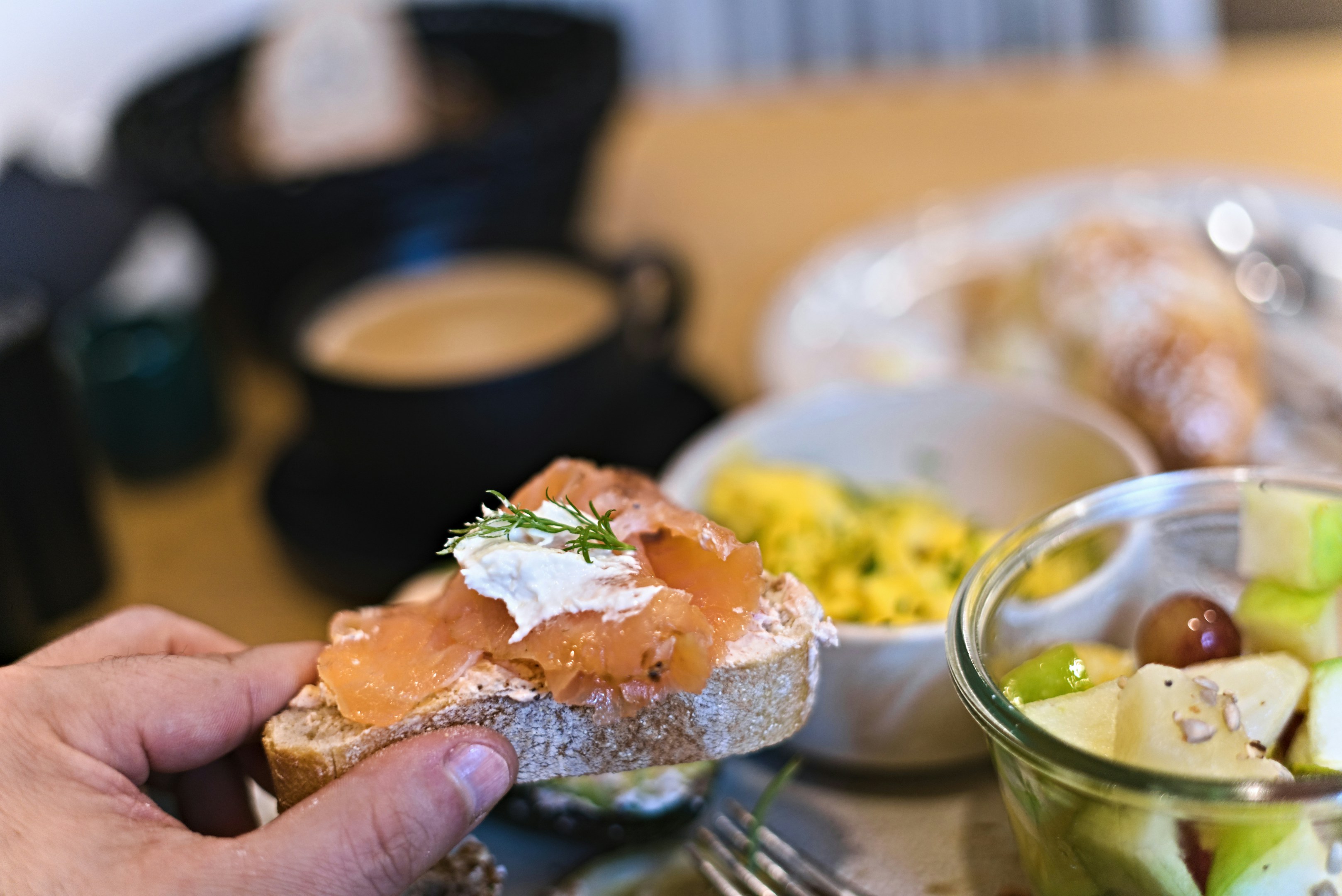 Hand holding salmon toast with fruit salad and coffee.