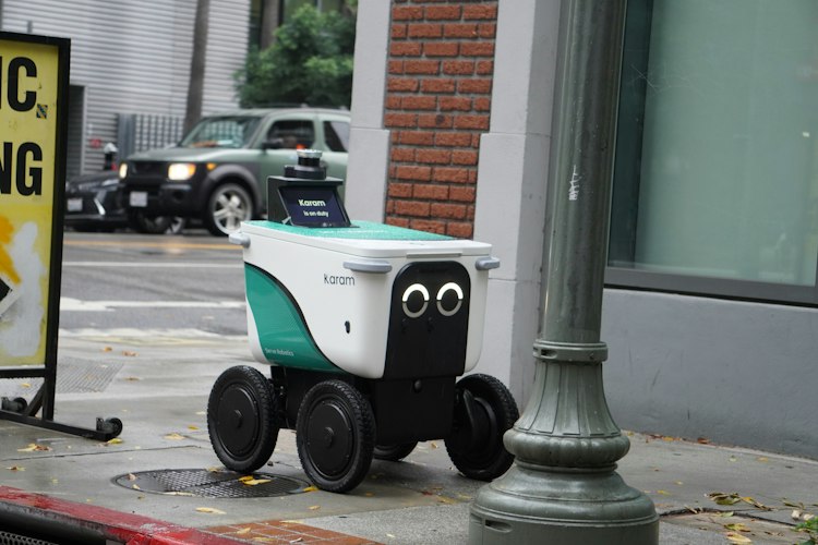 A small wheeled delivery robot rolling along a city sidewalk past pedestrians