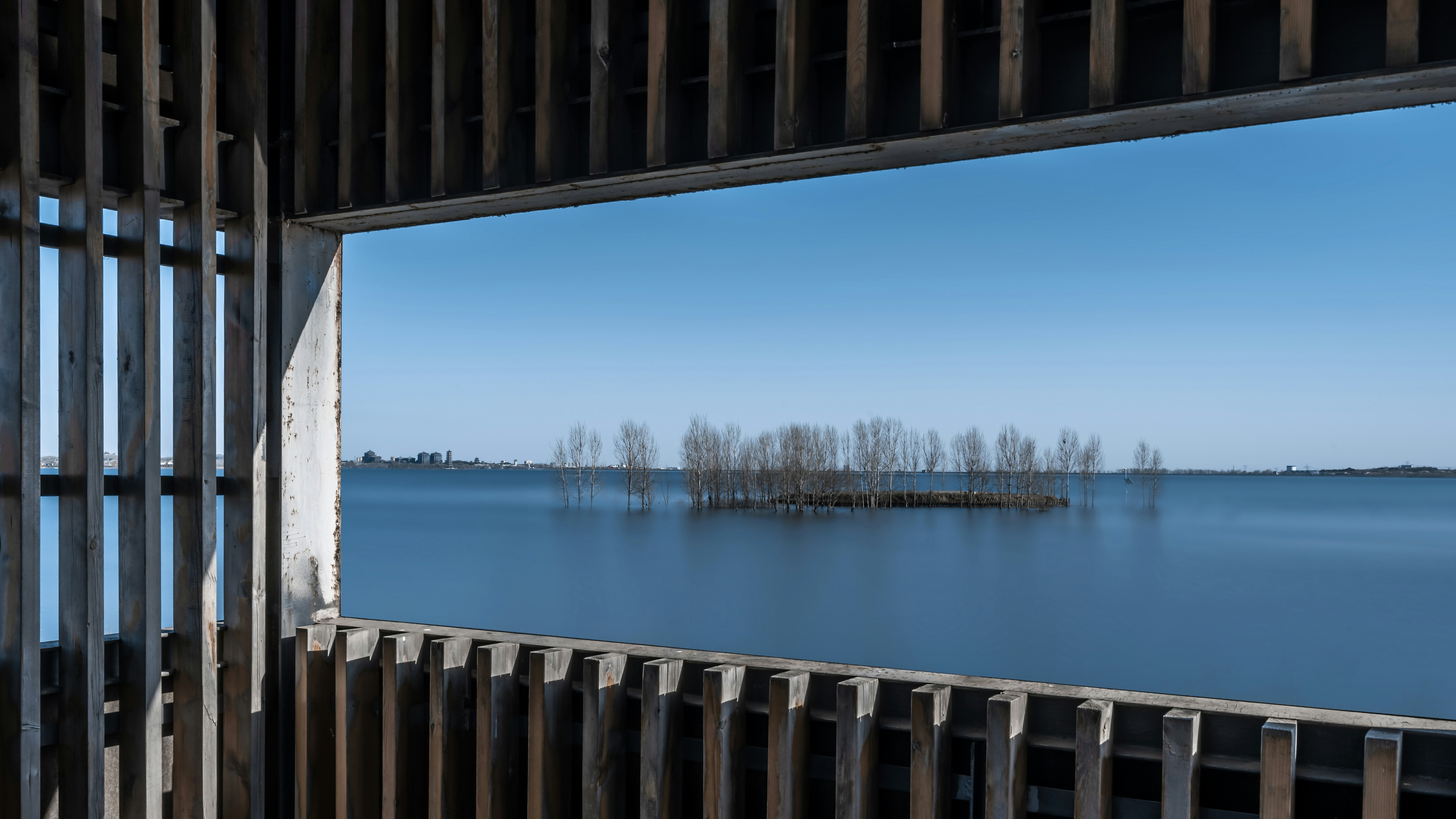 Blick durch die Holzkonstruktion mit ruhigem Wasser und entfernten Bäumen.