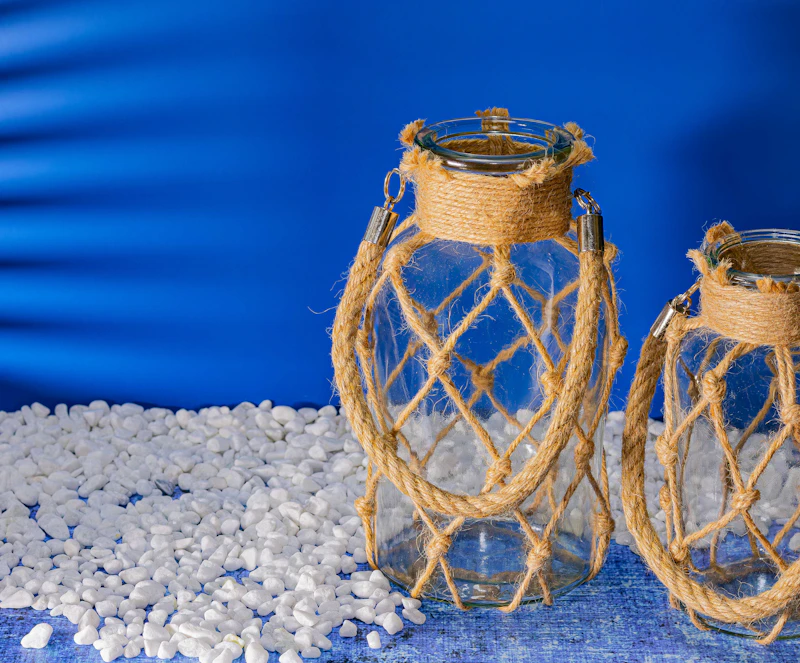 Two glass lanterns with rope accents on white pebbles