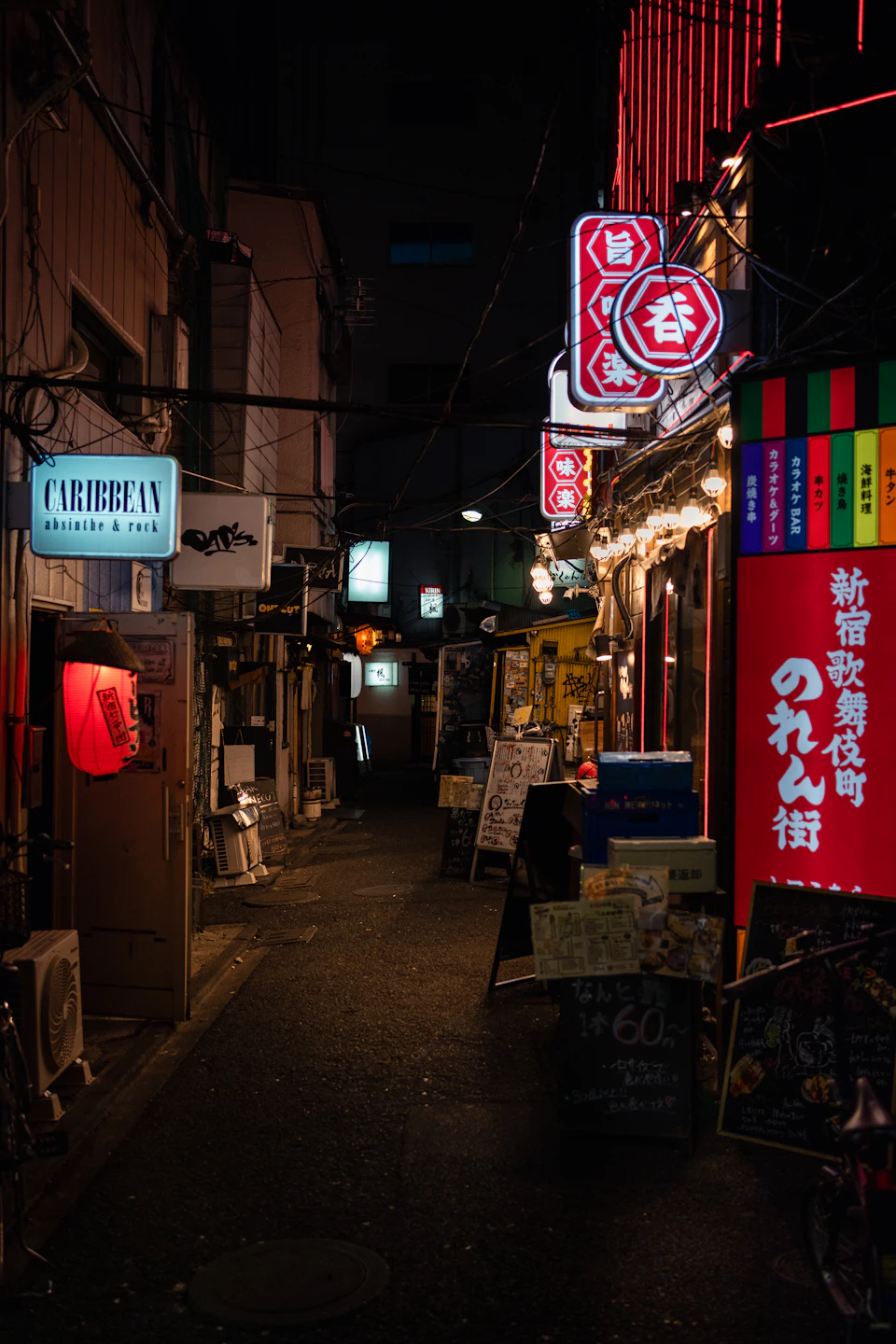 The image depicts a narrow alleyway in Japan at night, illuminated by neon signs and streetlights. The alley is lined with various shops and restaurants, including a sushi restaurant and a clothing store. A red sign stands out among the other signs, drawing attention to its presence. The perspective of the photo suggests it was taken from within the alley, providing a glimpse into the vibrant nightlife scene in Japan.