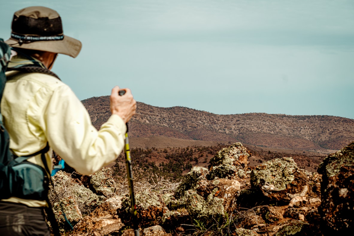 An older hiker with trekking poles standing on a high rocky ridge surveying the valley below, alone and at home