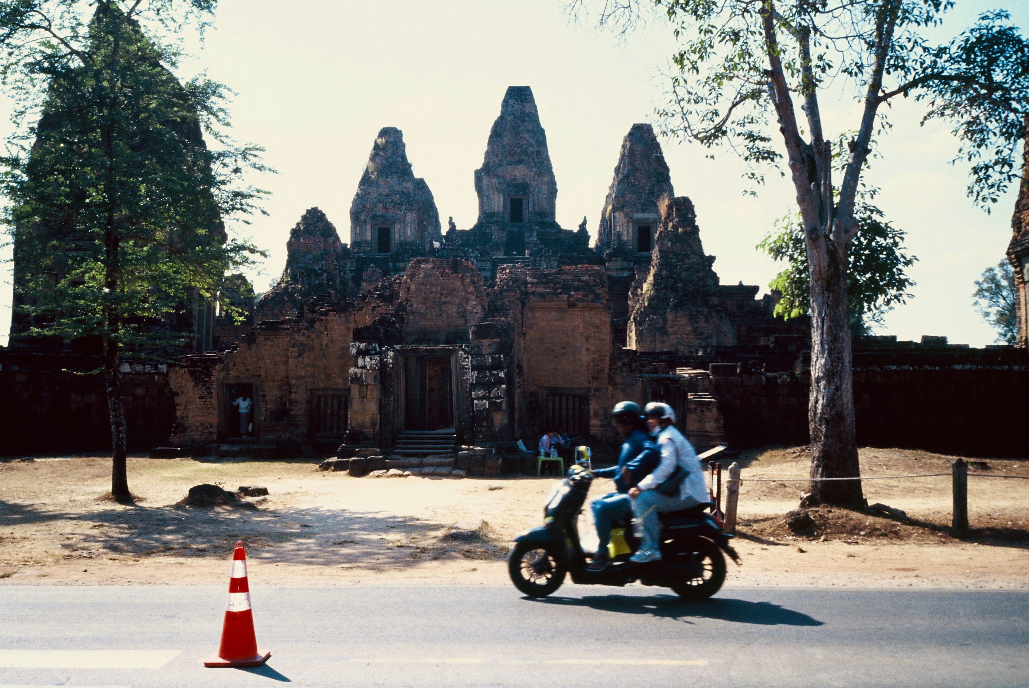 Zwei Personen fahren mit einem Roller an antiken Ruinen vorbei.