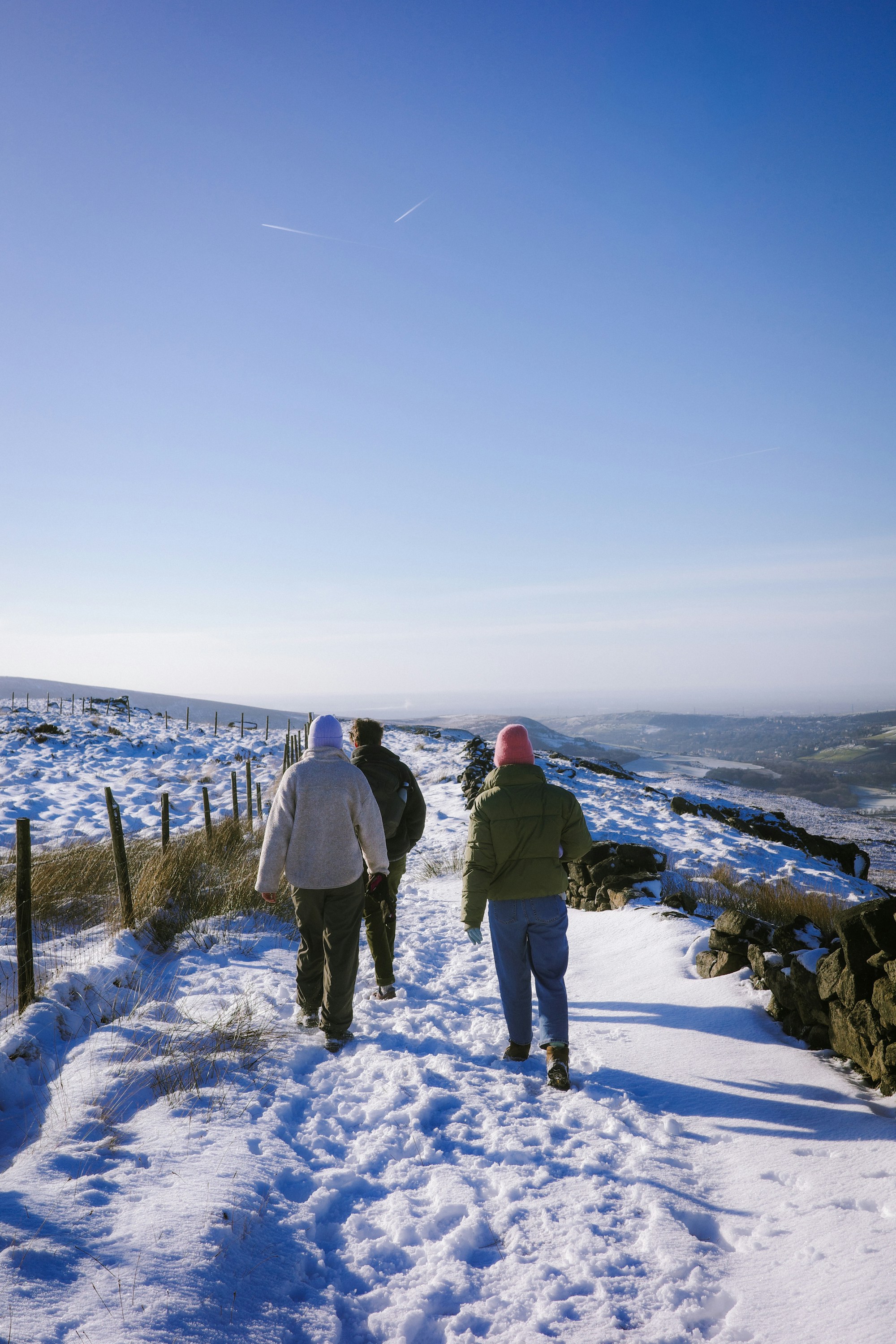 Three people walk on a snowy path overlooking hills.