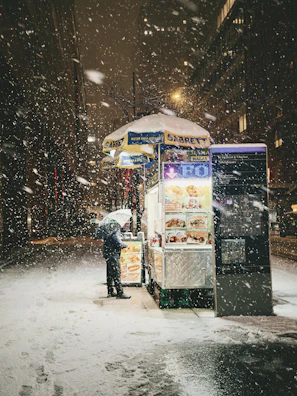 Person with umbrella at hot dog stand in snow.