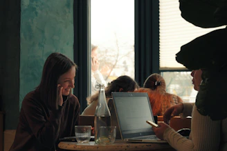 Two women talk at a table with a laptop.