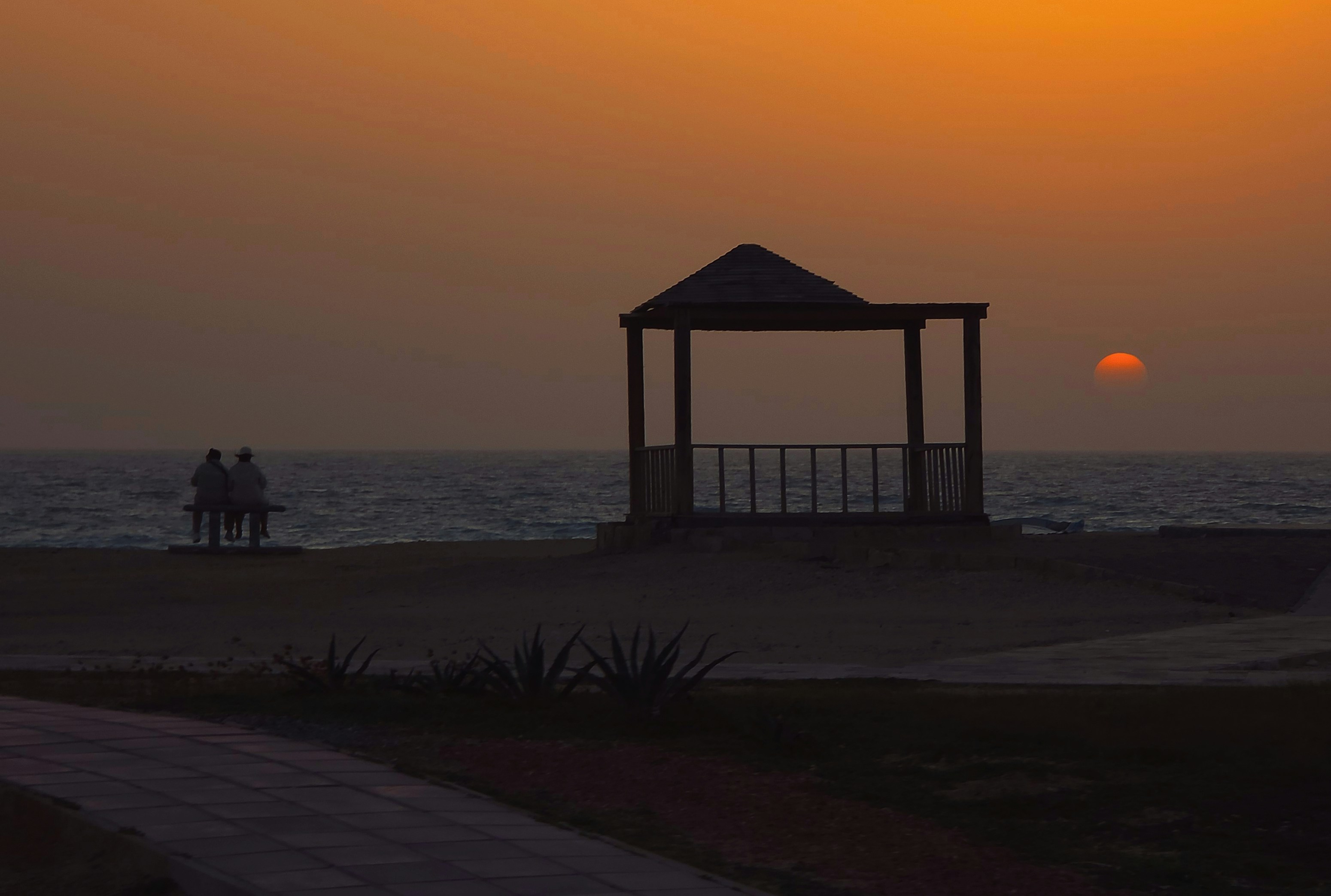 Two people watch sunset by a gazebo on the beach.