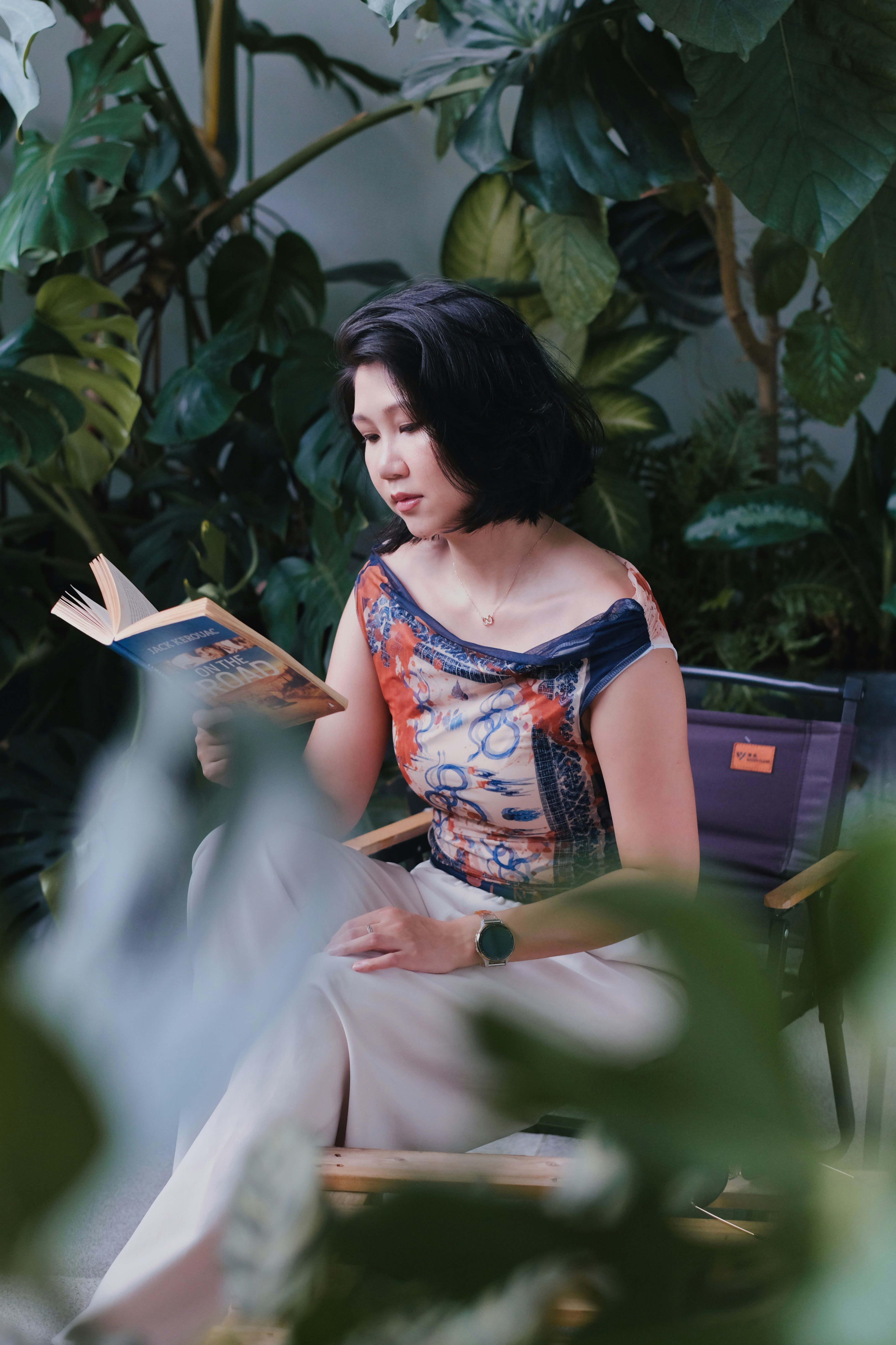 A woman reads a book surrounded by lush green plants.