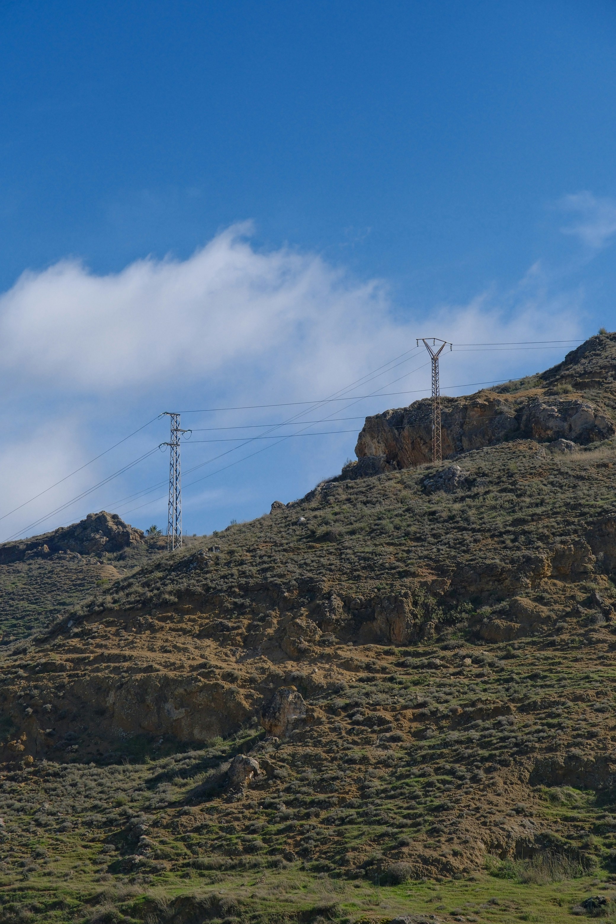 Power lines cross a grassy, rocky hillside under blue sky.