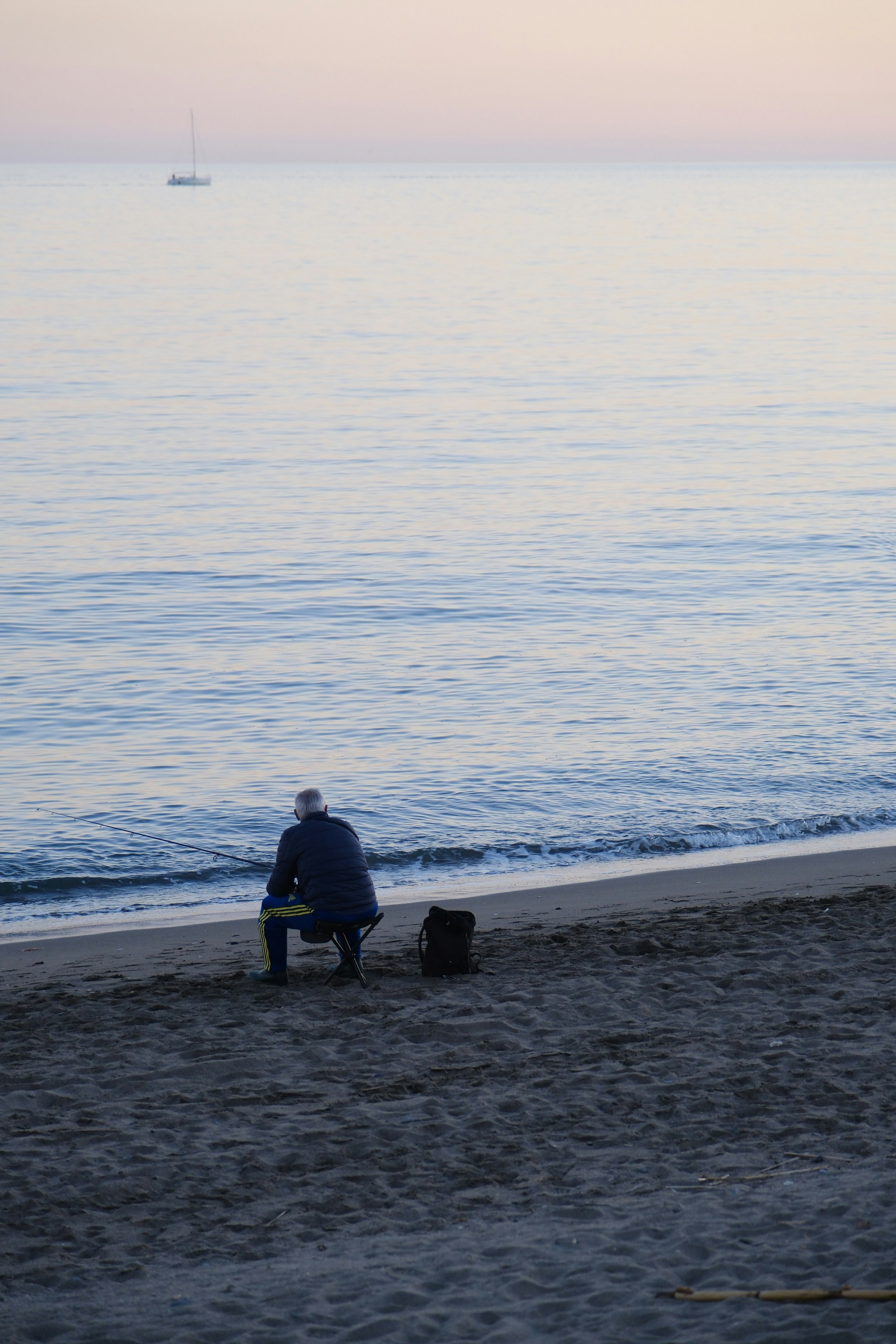 Man sits on beach watching sailboat on calm ocean.