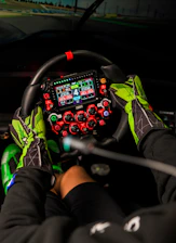 Hands in racing gloves grip a complex steering wheel.