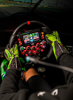Hands in racing gloves grip a complex steering wheel.