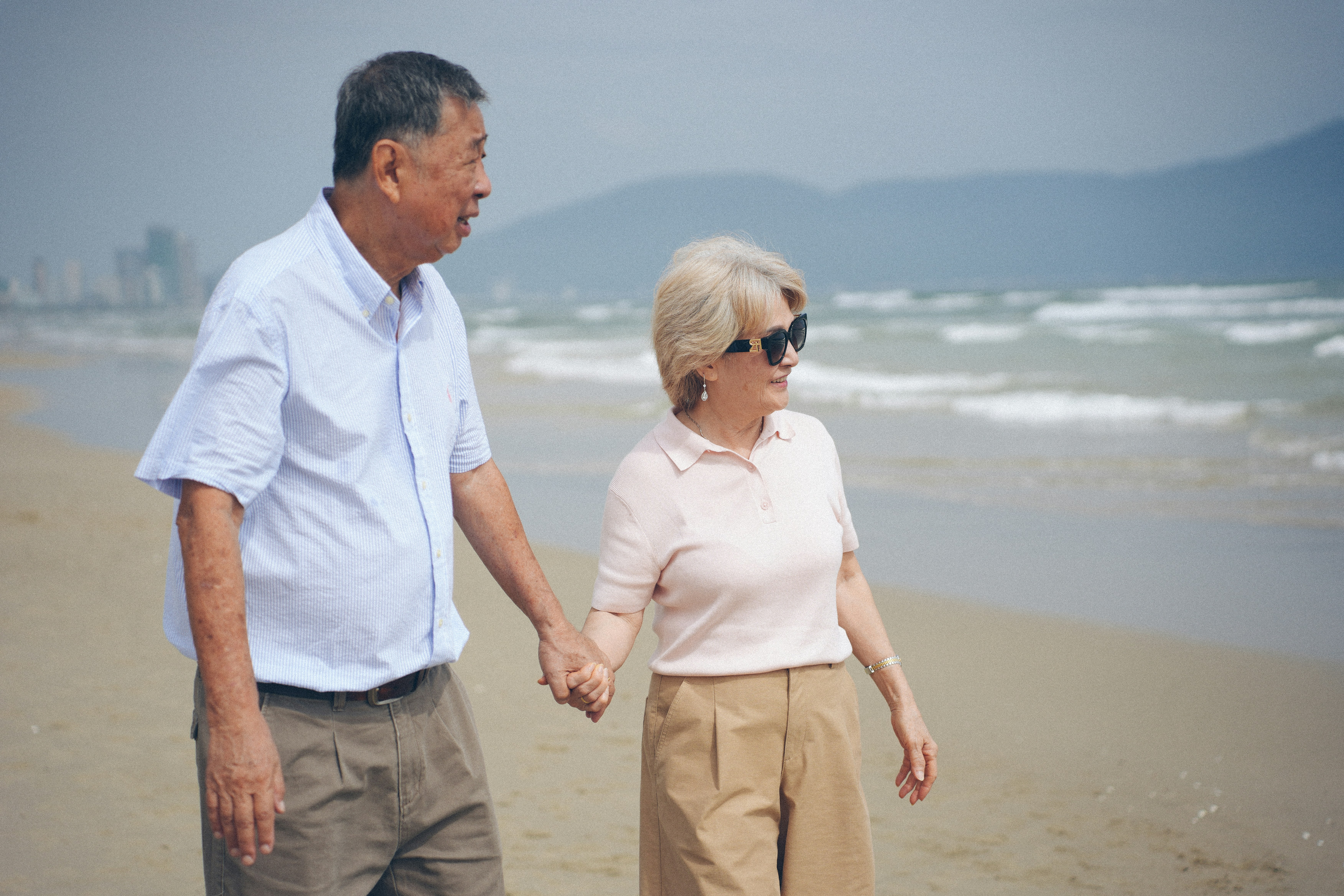 Elderly couple holding hands walking on a sandy beach.