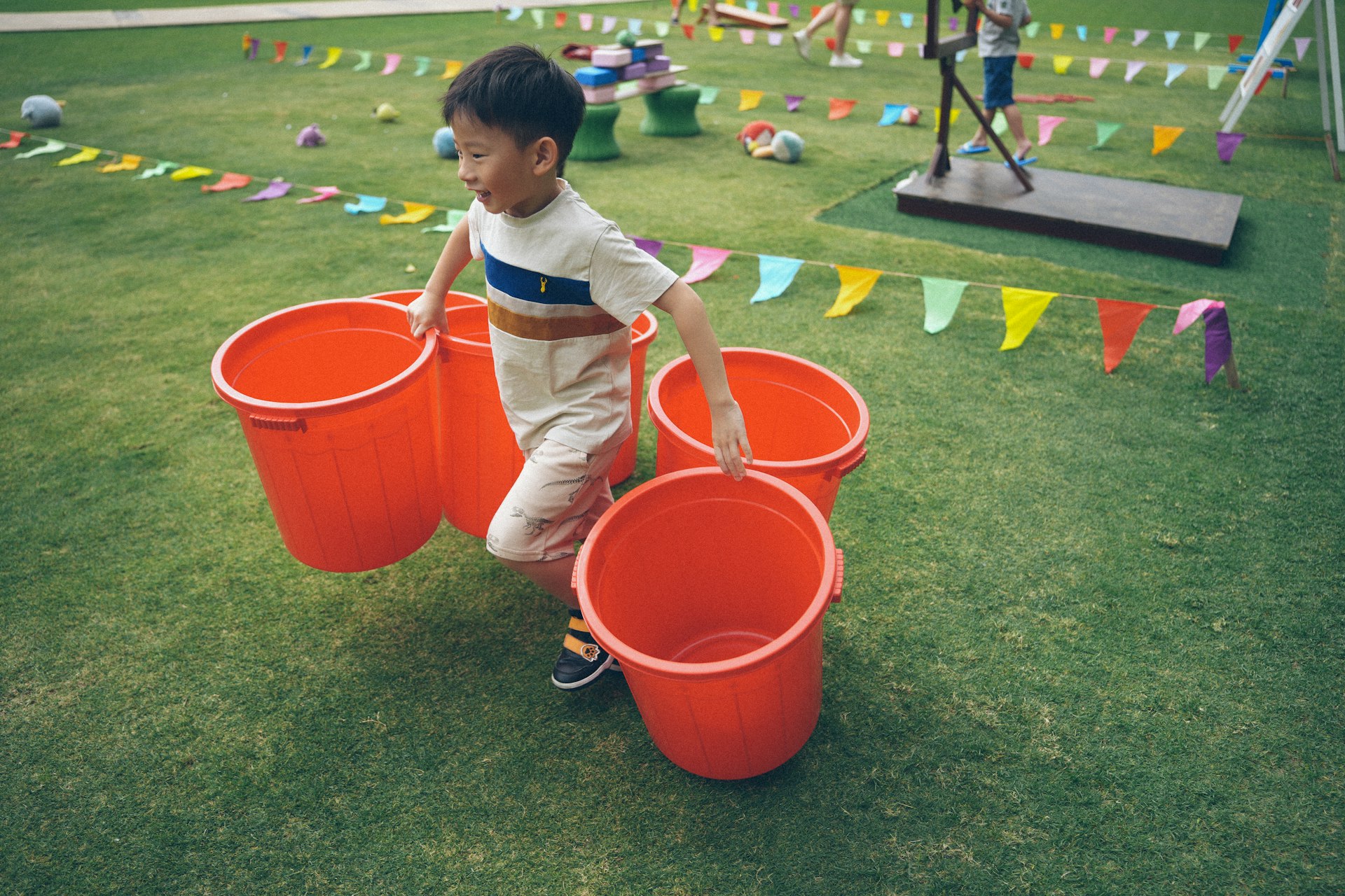 A young boy carries several large red buckets on grass.