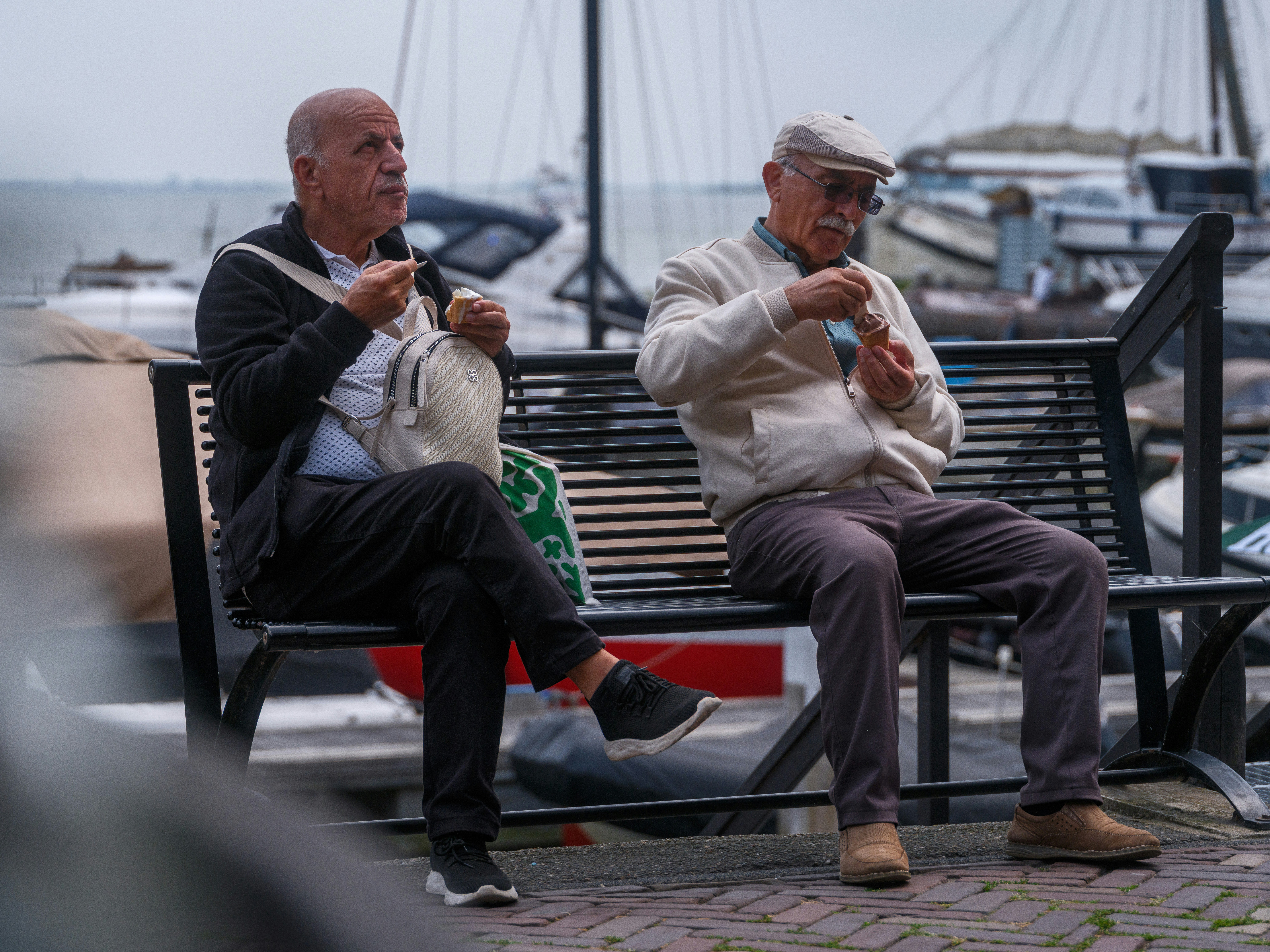 two men eating ice cream while sitting on a park bench