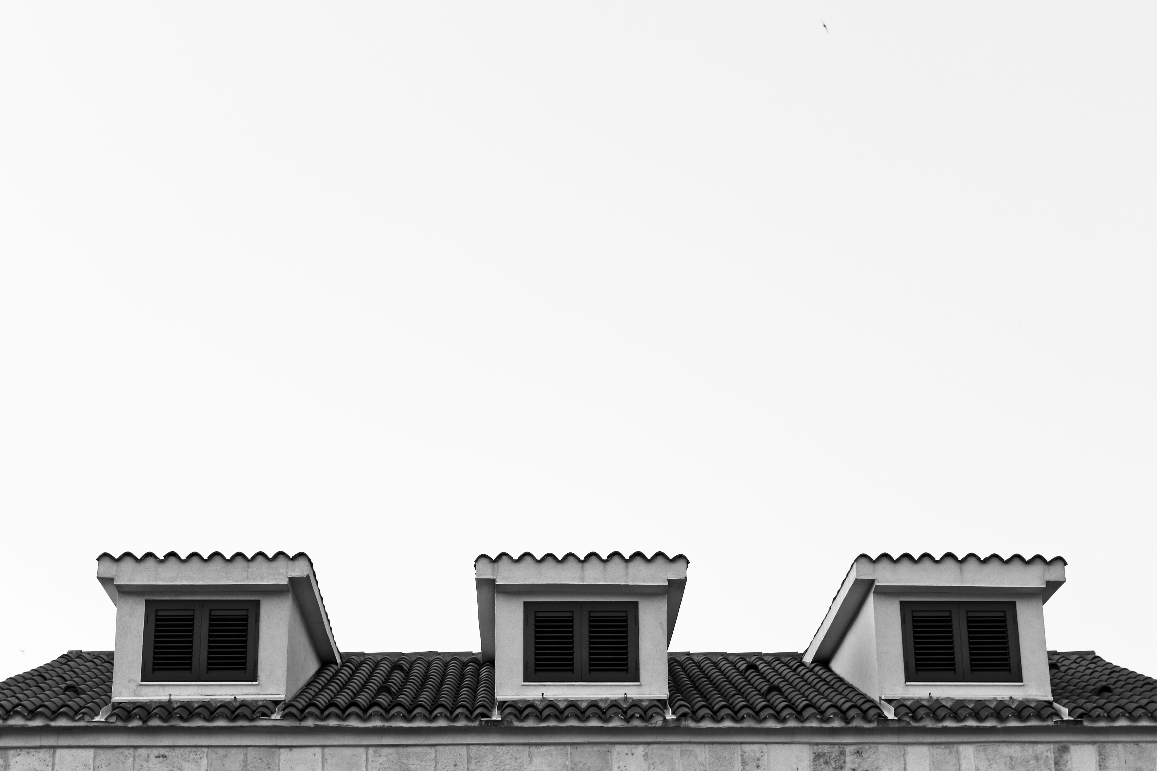 Three symmetrical dormer windows with dark shutters sit atop a tiled roof against a vast, minimalist white sky