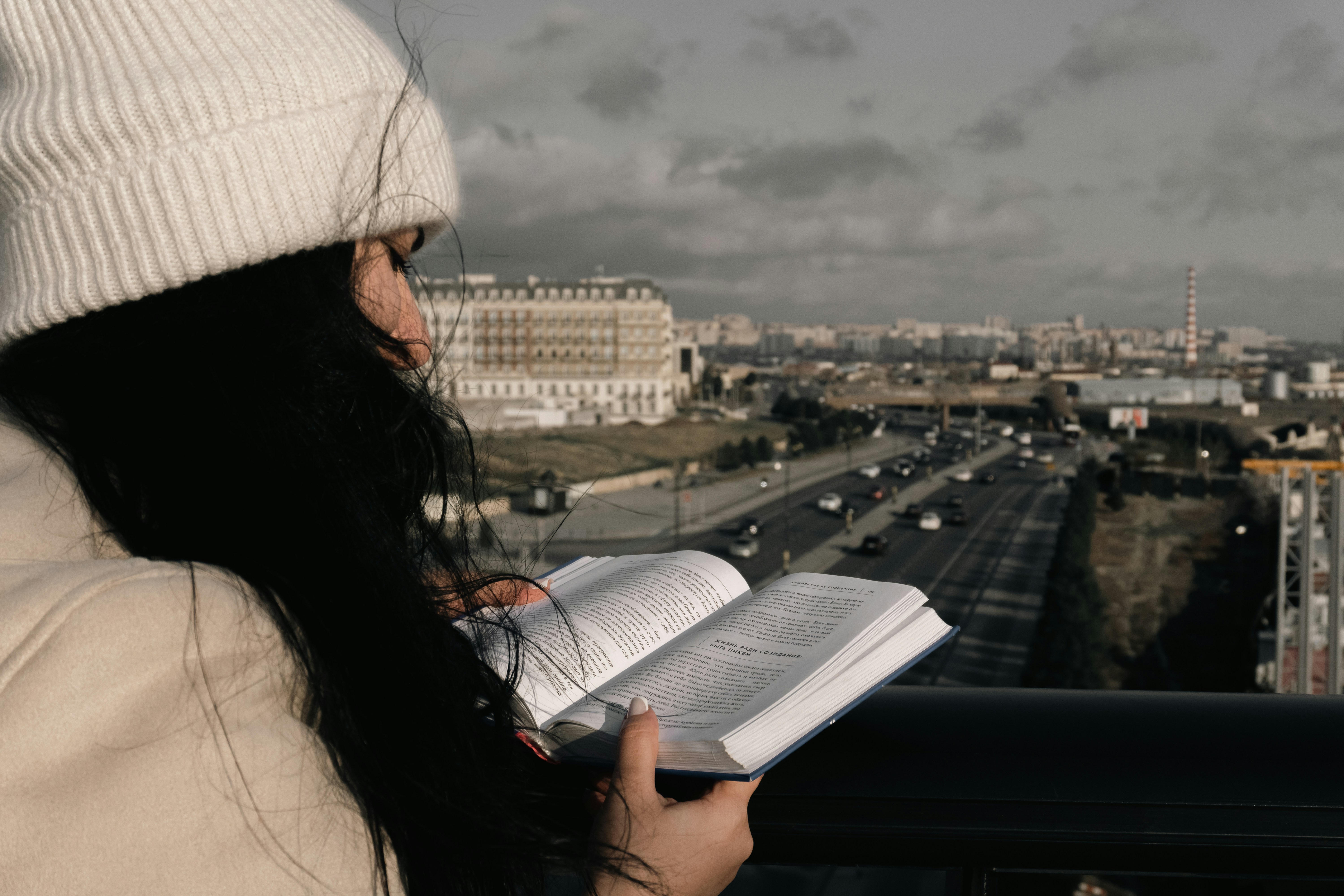 Woman in beanie reading book overlooking city highway
