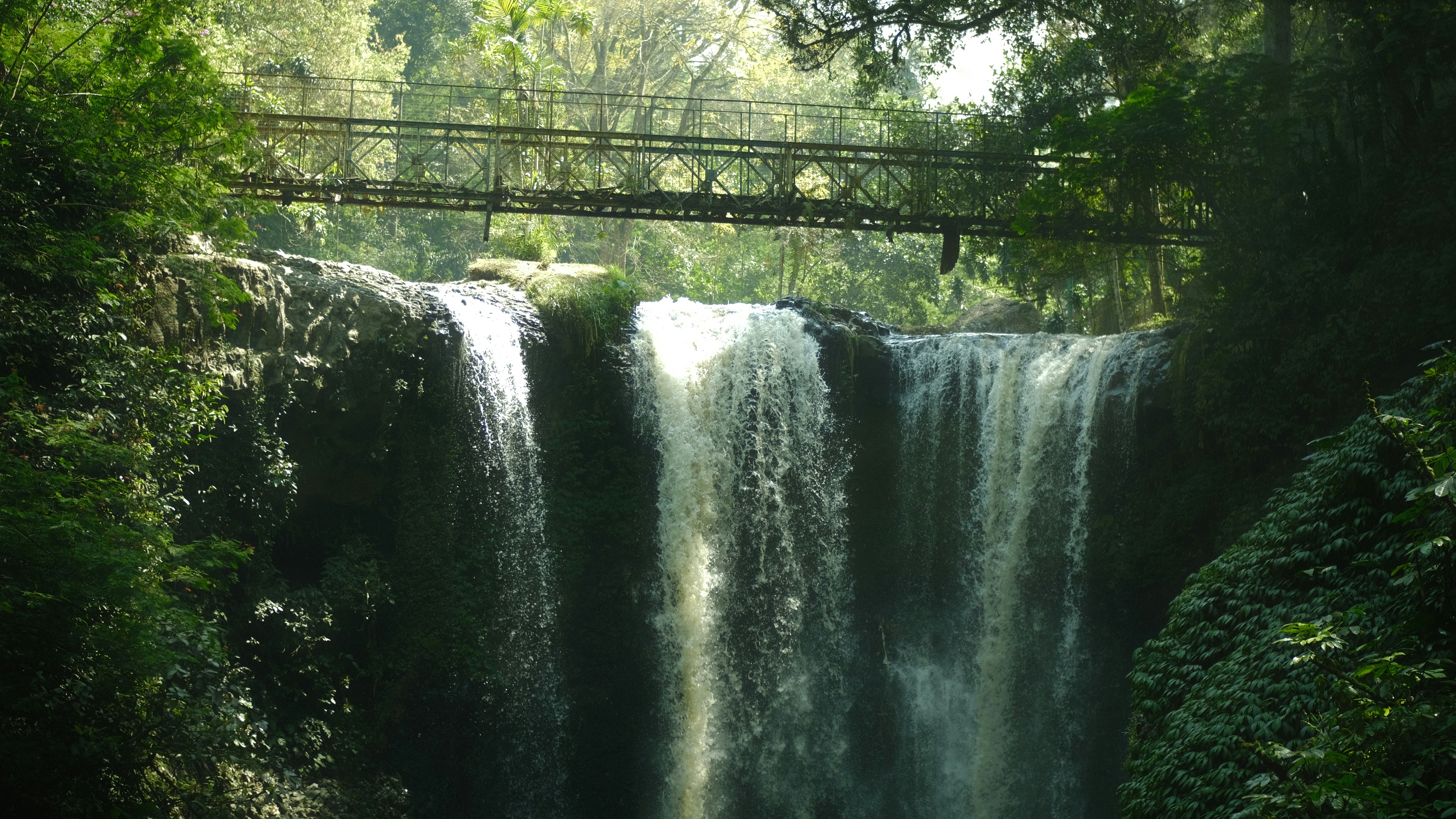Air terjun yang rimbun mengalir menuruni bebatuan dengan jembatan di atasnya.