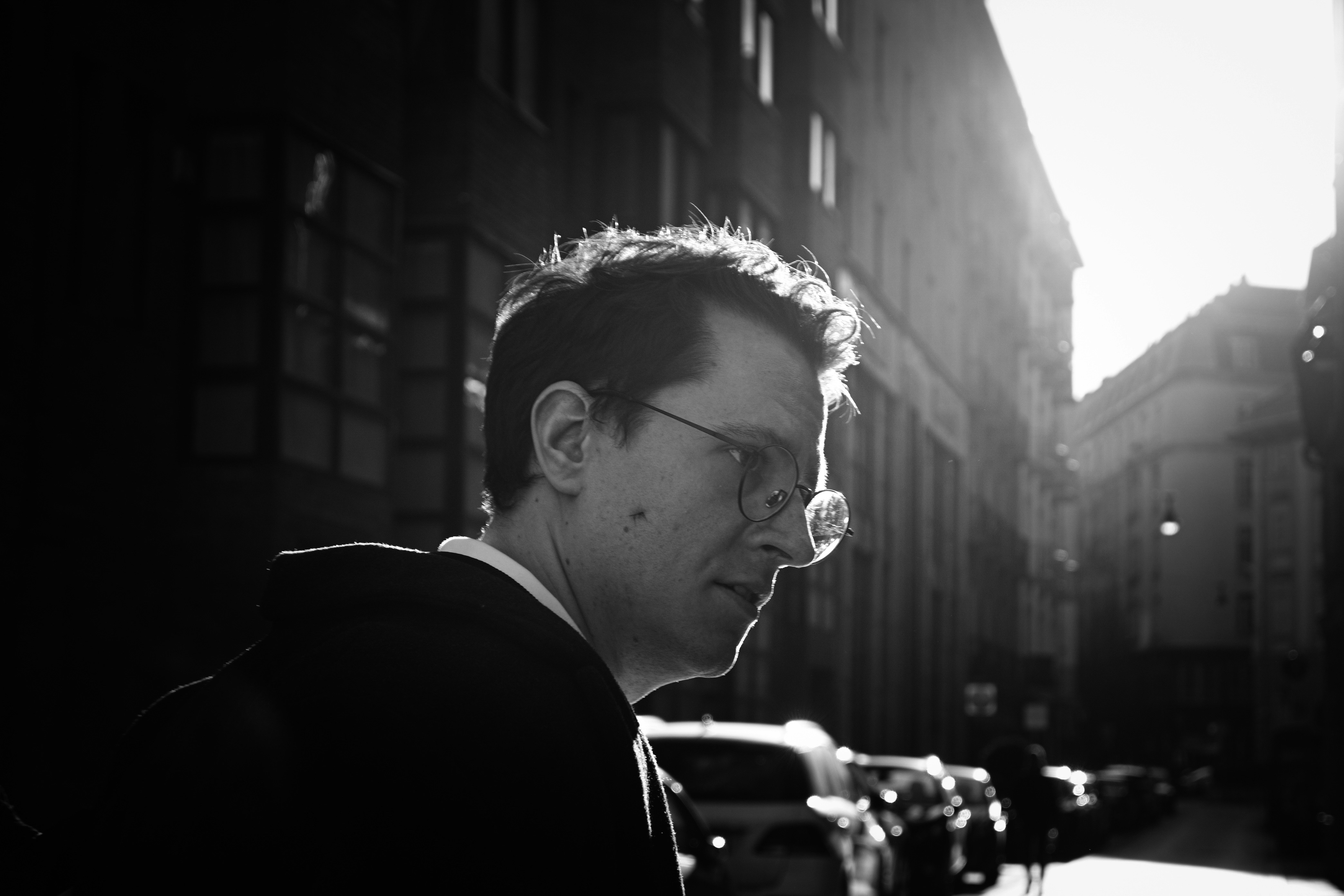 Man with glasses walks down a sunlit street.