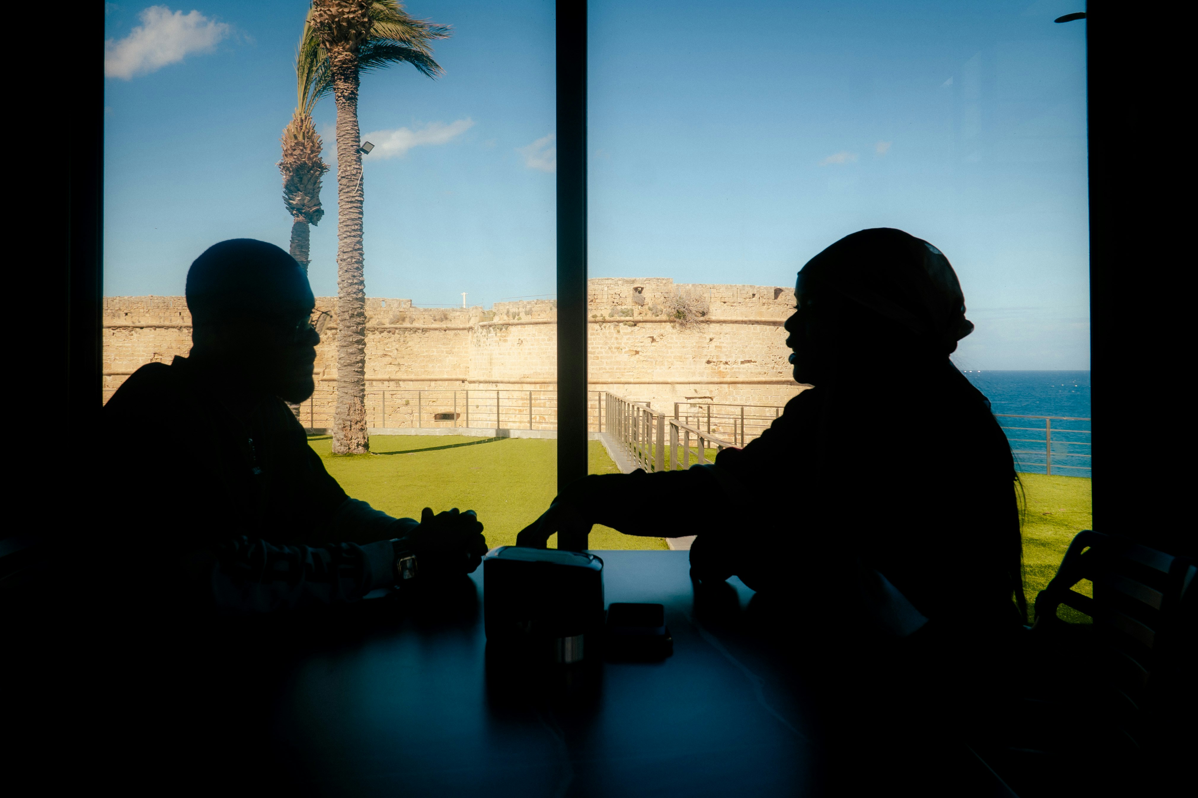 Dua orang bersiluet berbicara di meja di tepi laut.