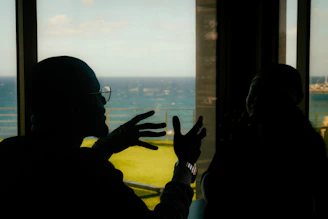 Two people silhouetted talking by a window overlooking ocean.