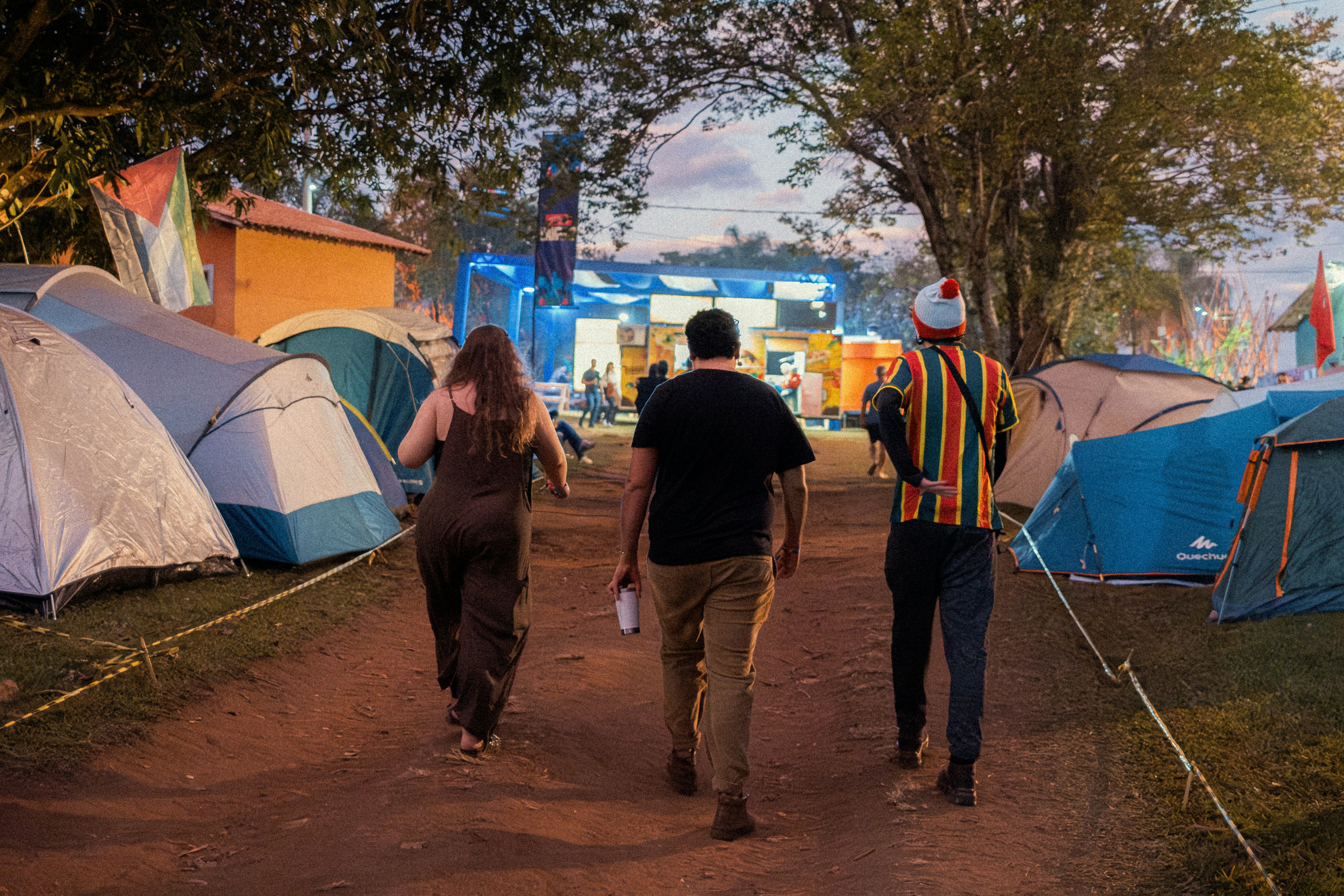 Three people walk through a tent camp at dusk.