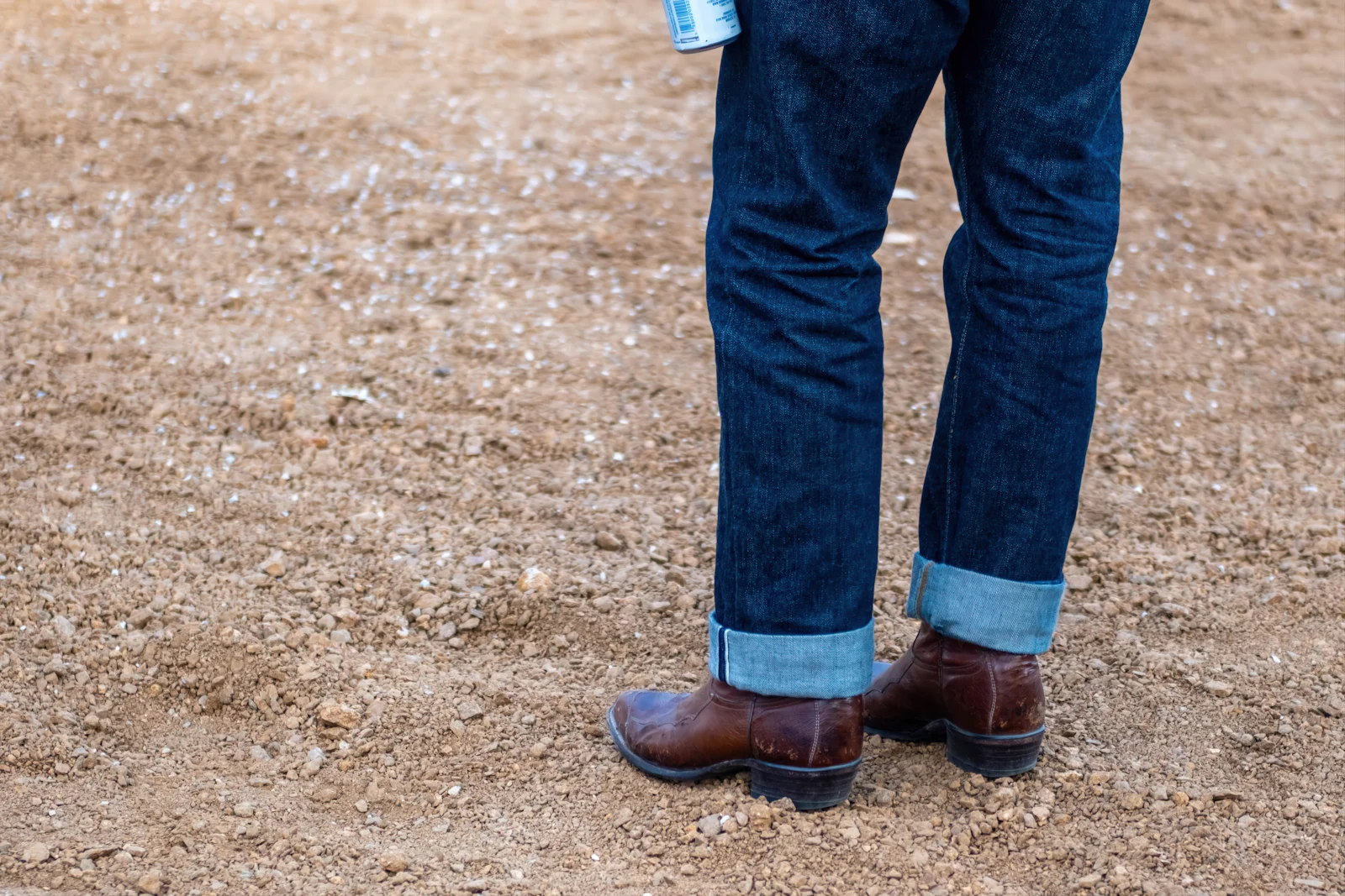 Cuffed denim over brown leather boots, standing on dirt.