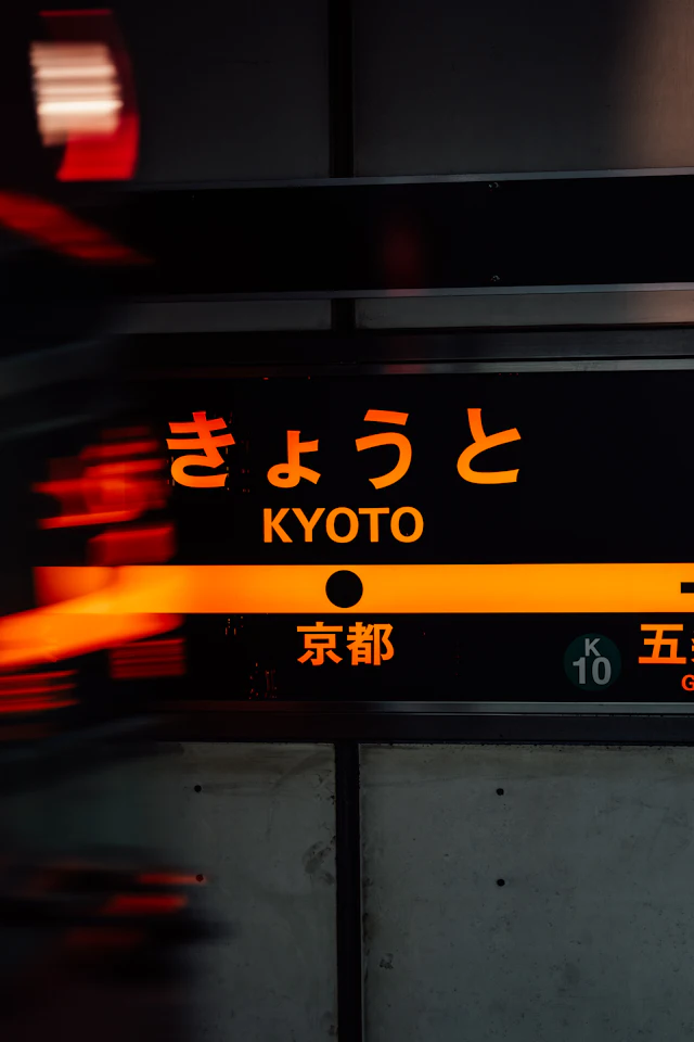 An illuminated platform sign reading 'Kyoto' in Japanese and English, blurred by a passing train.