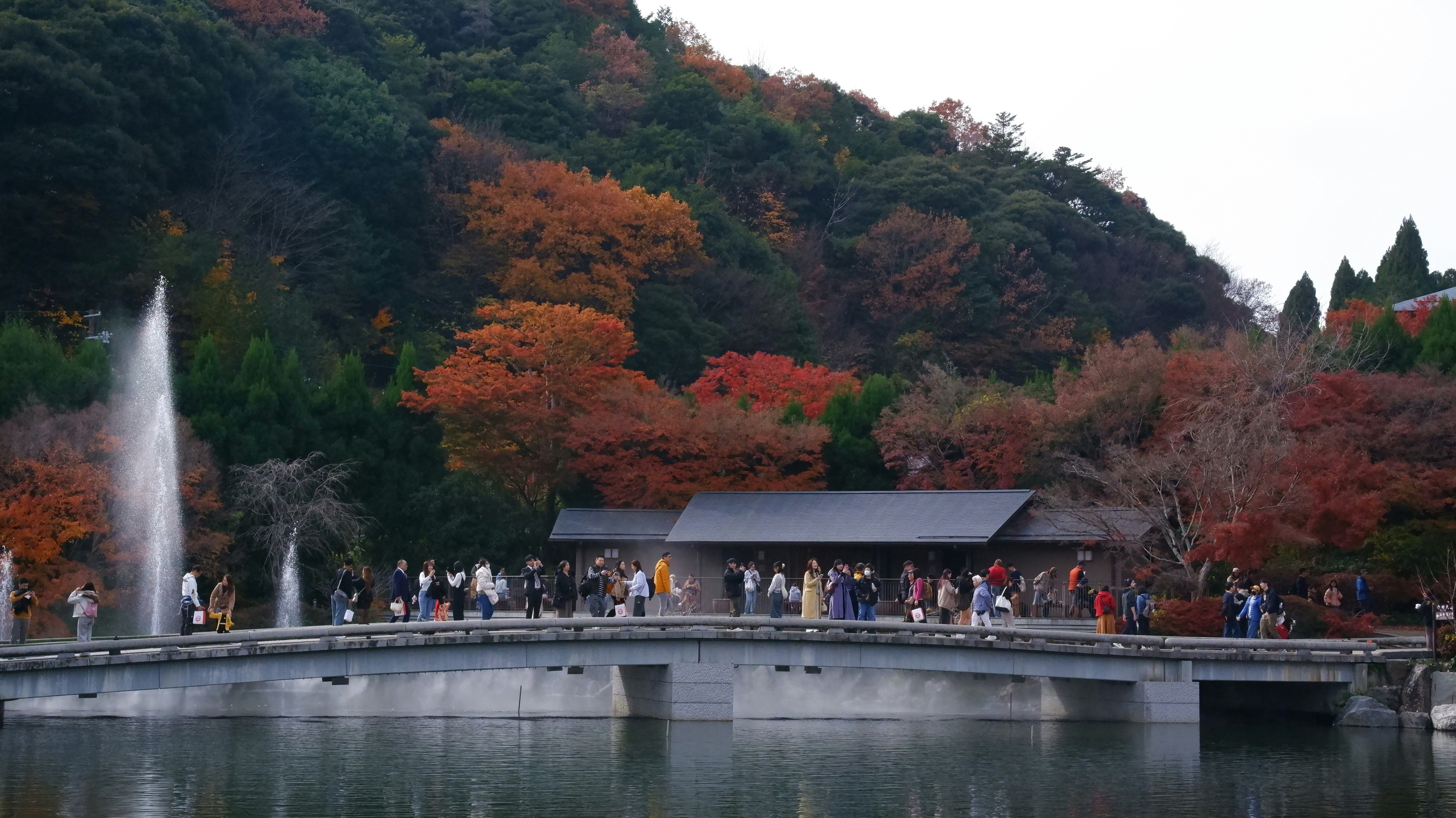 People walk across a bridge with autumn foliage and fountain.