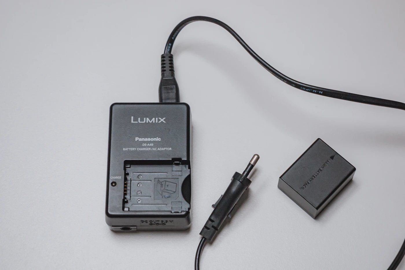 A camera battery charger plugged into a cable, with a rectangular lithium-ion camera battery lying beside it on a white surface