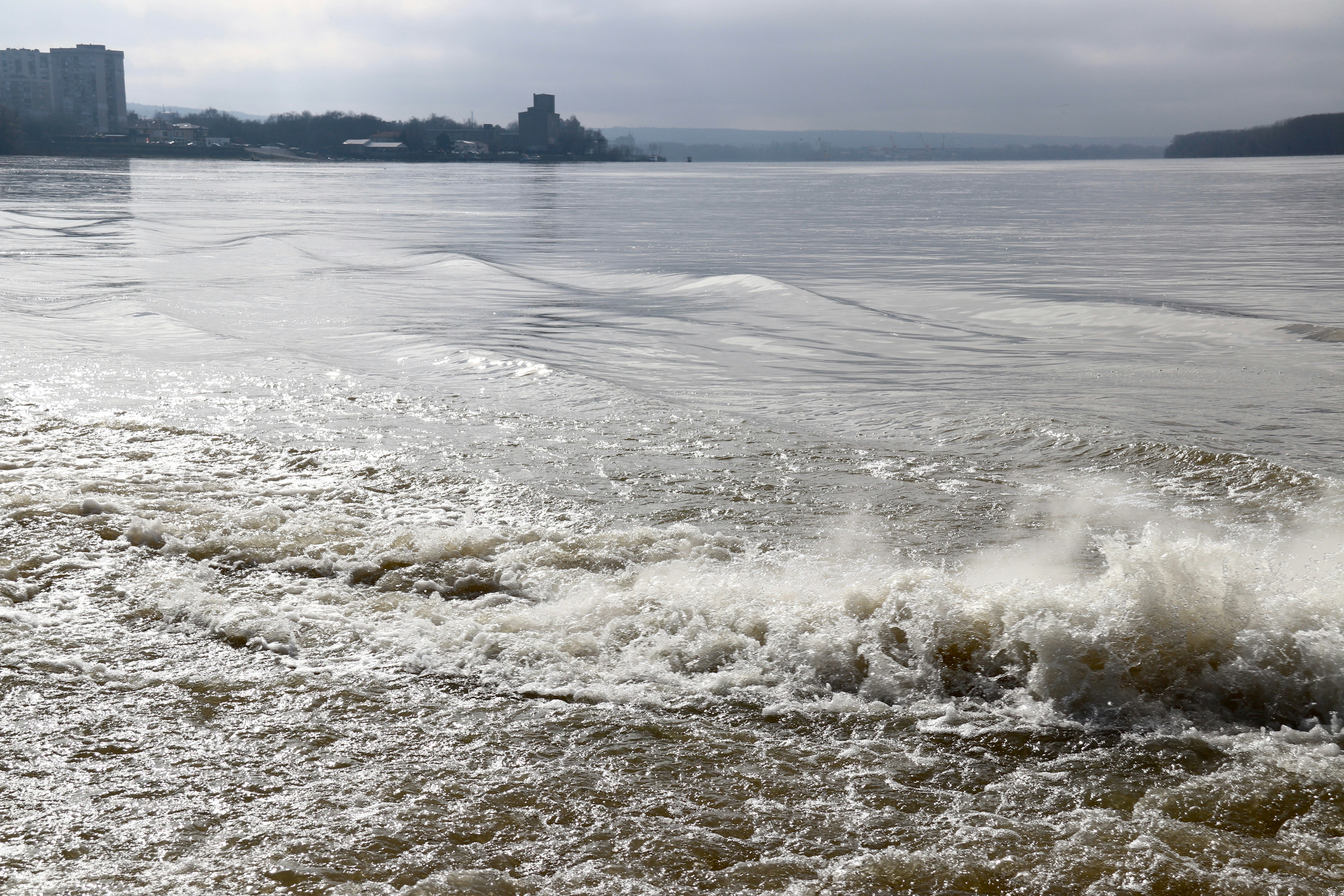Turbulent water flows past a distant cityscape under cloudy skies.