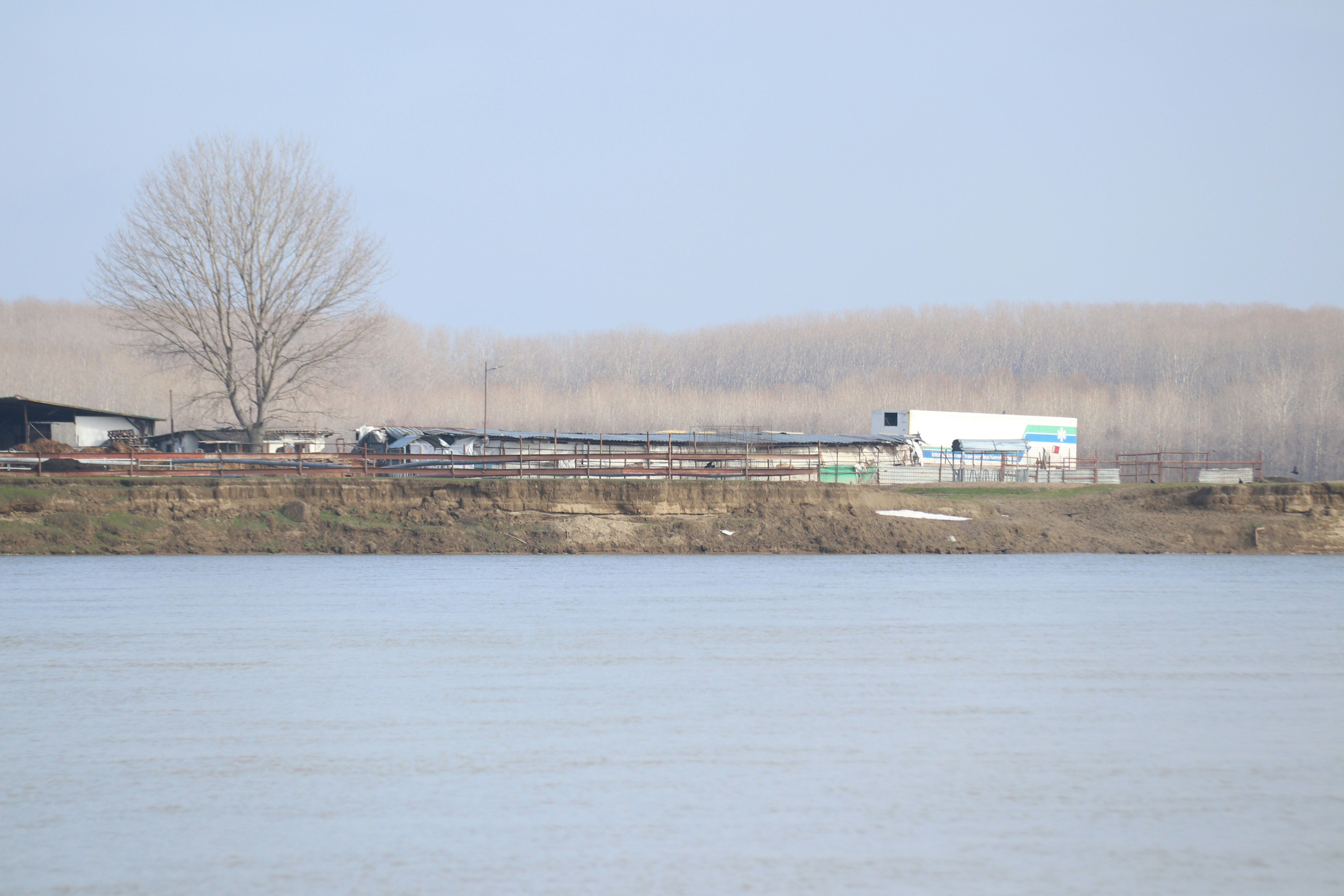 Edificios industriales en una orilla de río con árboles desnudos.