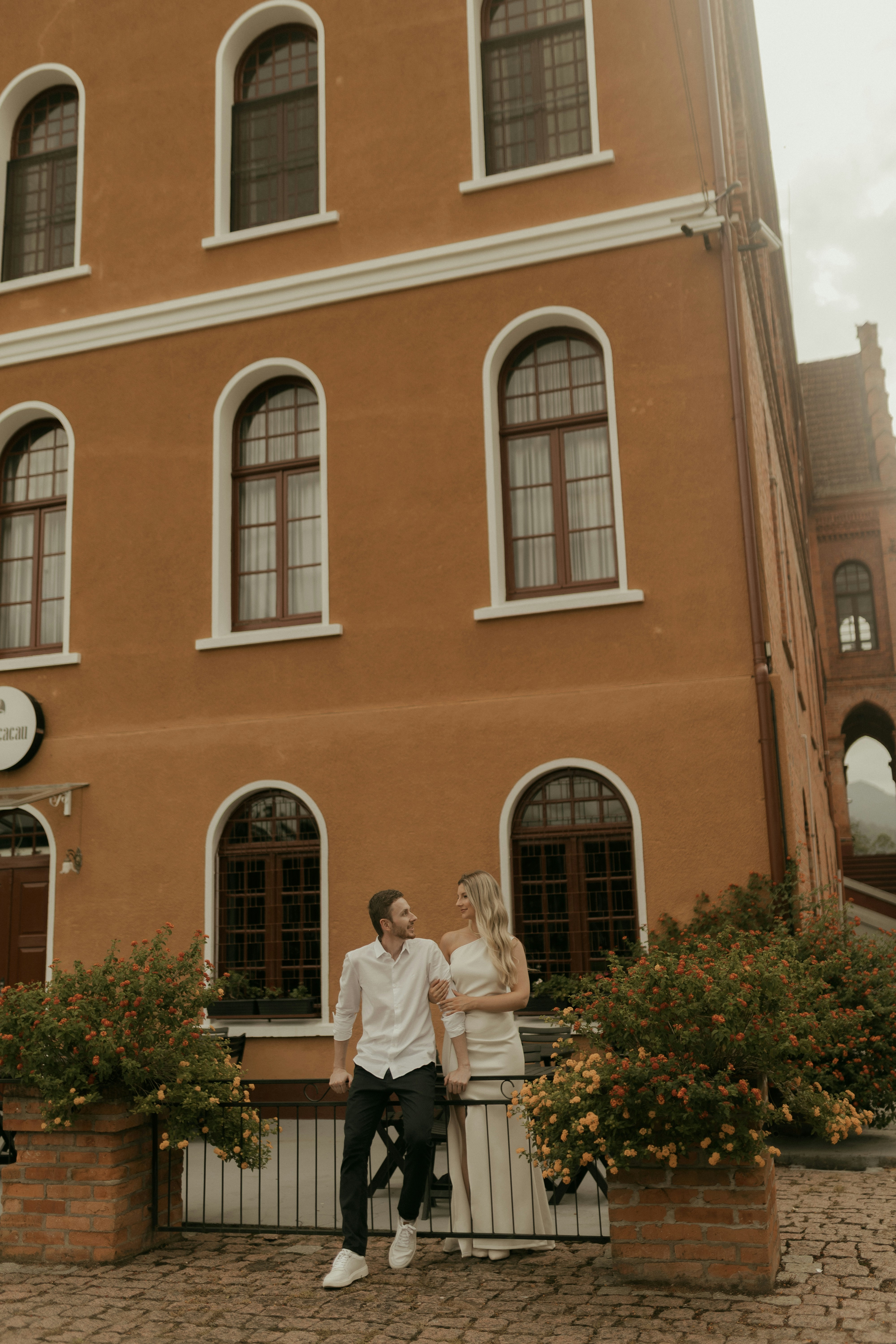 A couple stands outside a building with arched windows.
